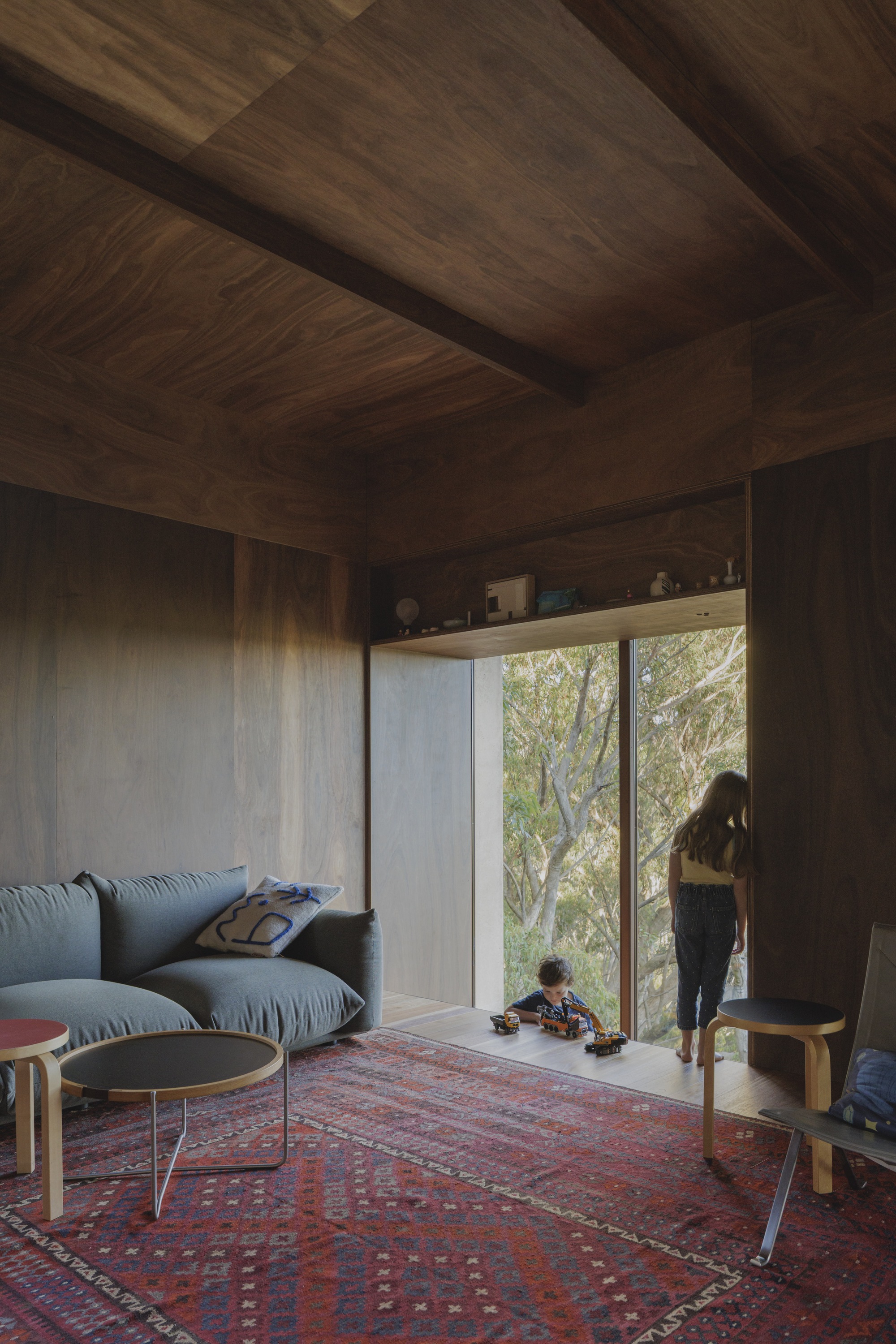 Walnut-panelled sitting area with a deep window seat where a child plays with toy trucks and a girl looks out at the trees