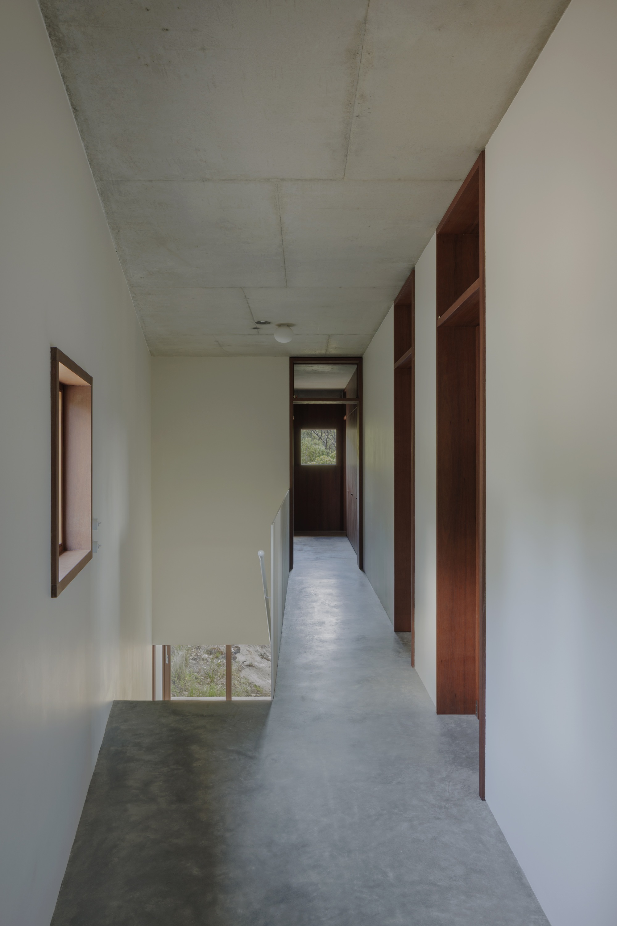 Upper hallway with polished concrete floor, timber-framed wardrobe niches and a small window looking onto a stairwell and bushland beyond