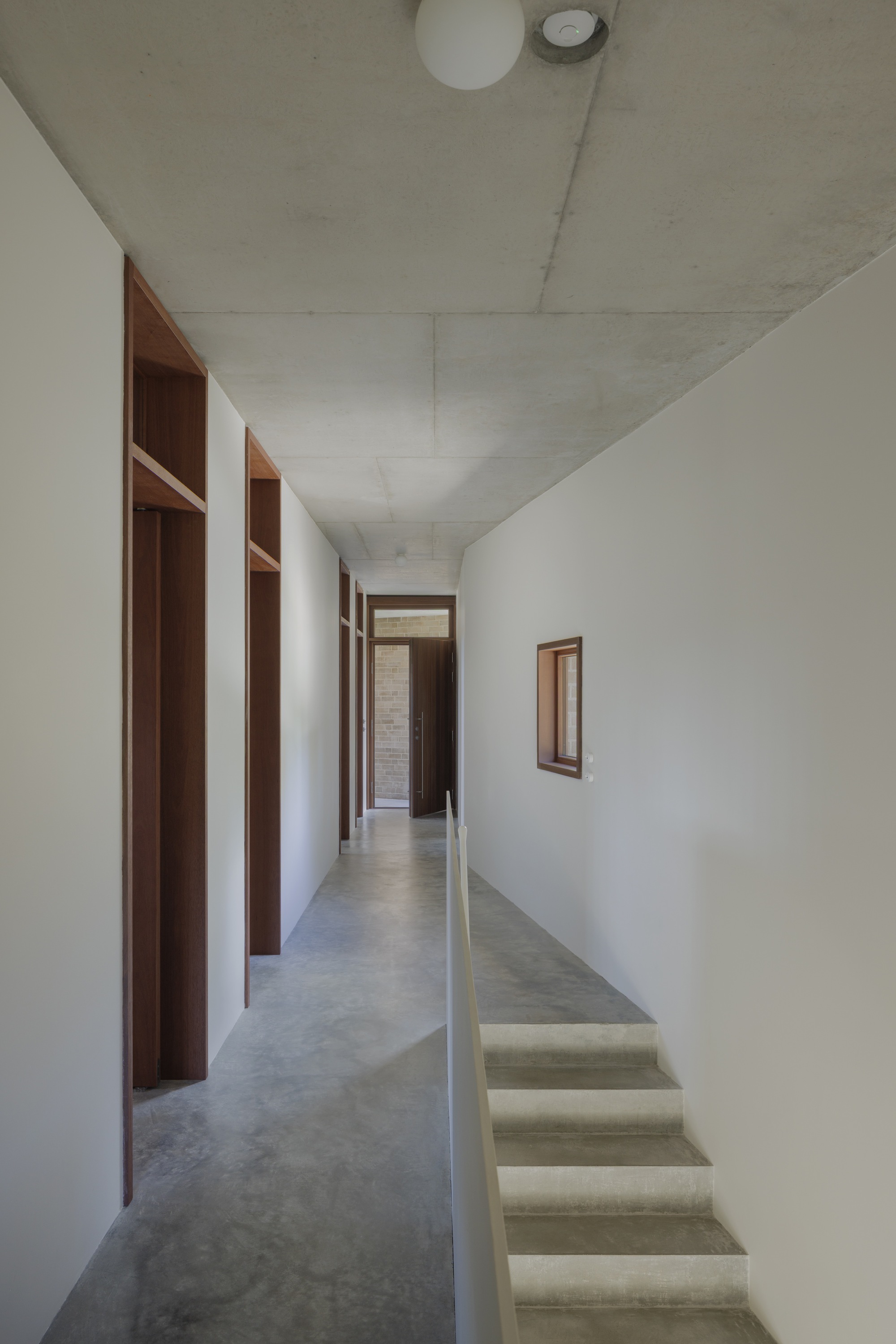 Corridor with concrete stair descending beside a white wall, timber door frames and a small square window in soft daylight