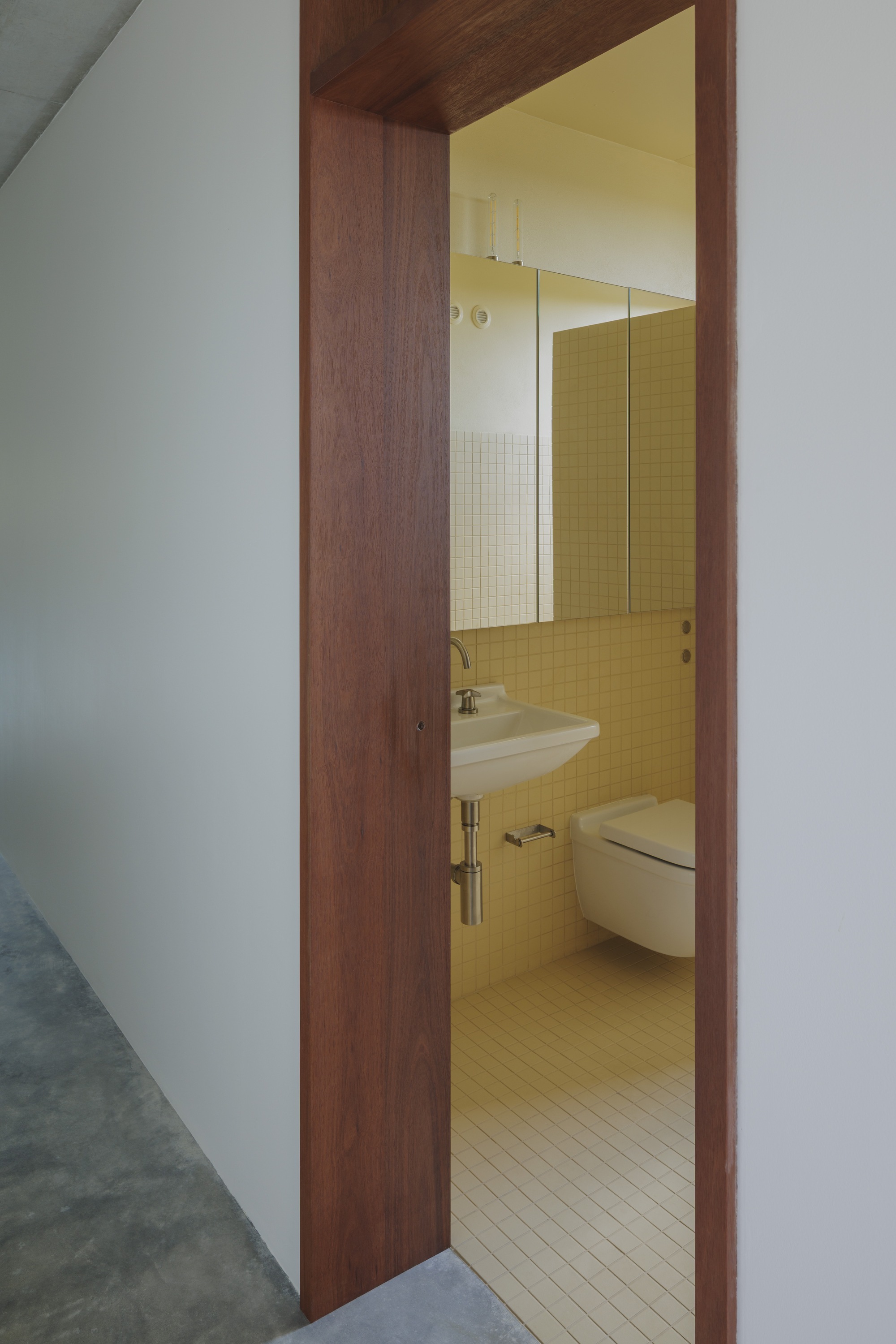 Timber door frame opening onto a bathroom with yellow square tiles, wall-hung basin and mirrored cabinet in soft light