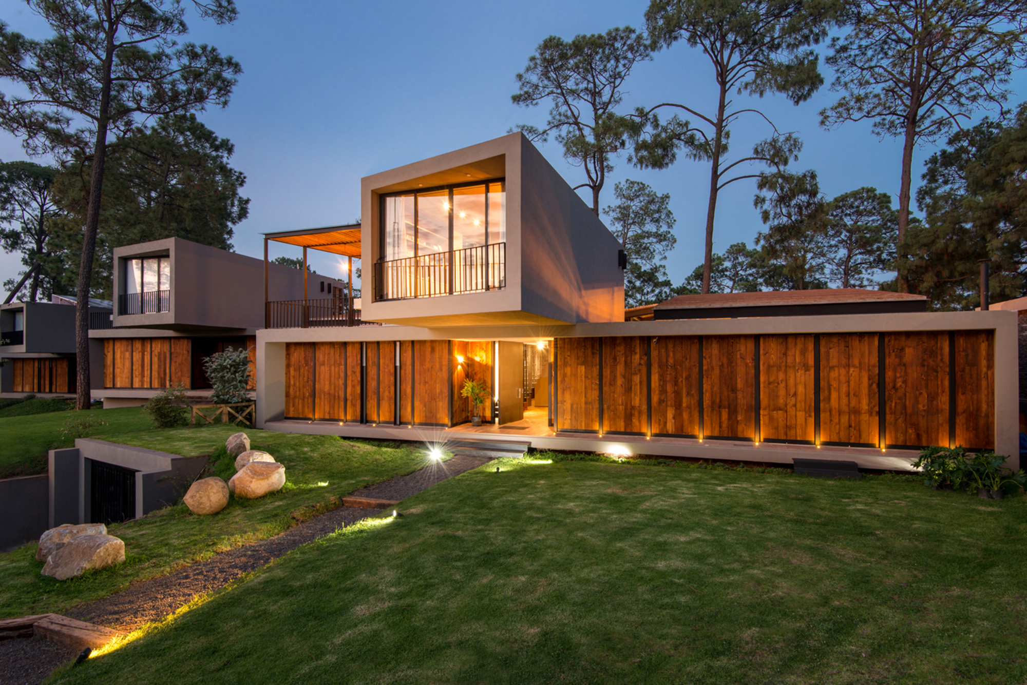 Two-storey concrete volumes with timber slat panels lit by ground spotlights on a lawn among pines at dusk