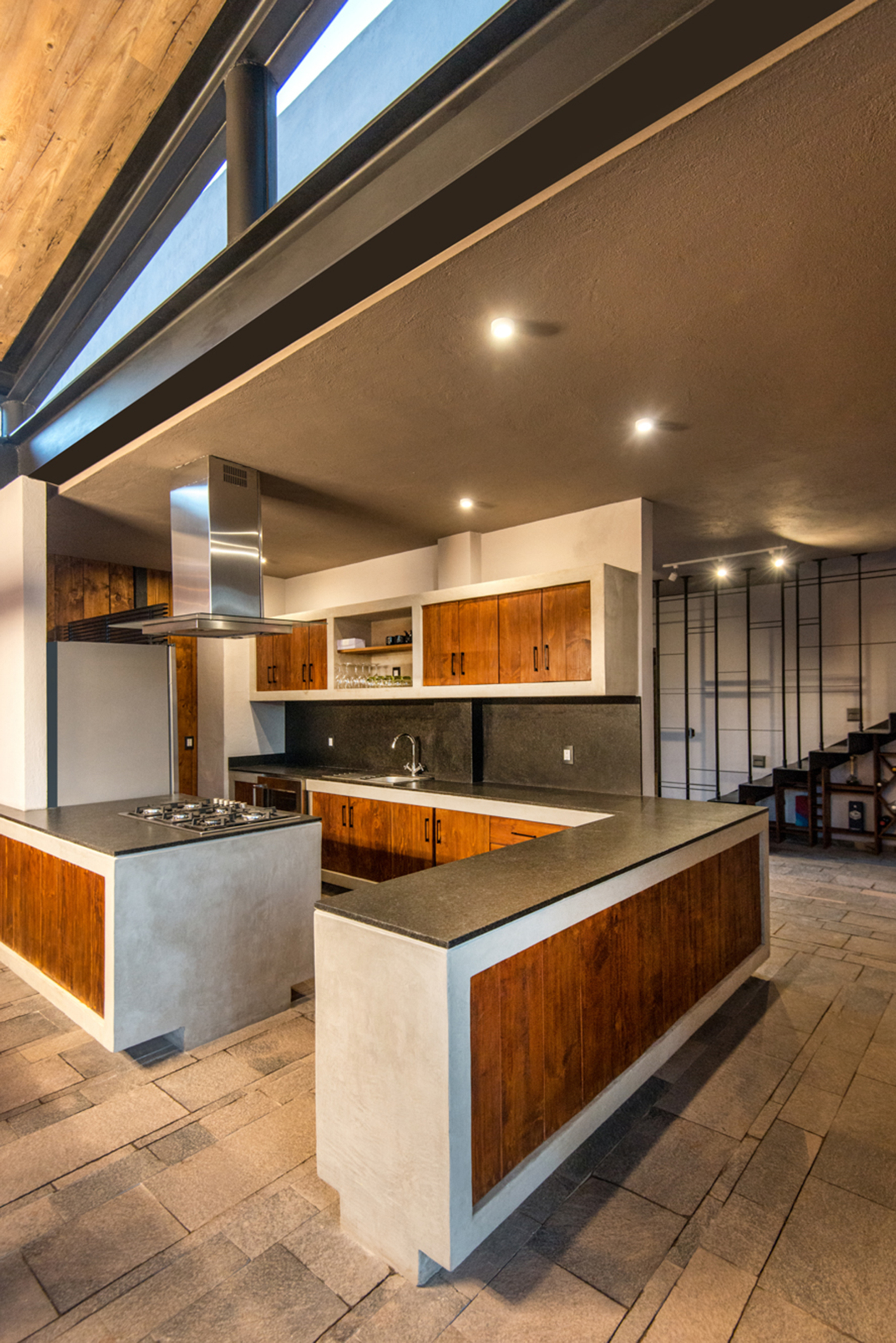Kitchen with concrete and timber-clad islands, dark stone counters and steel stair behind, lit by a clerestory band