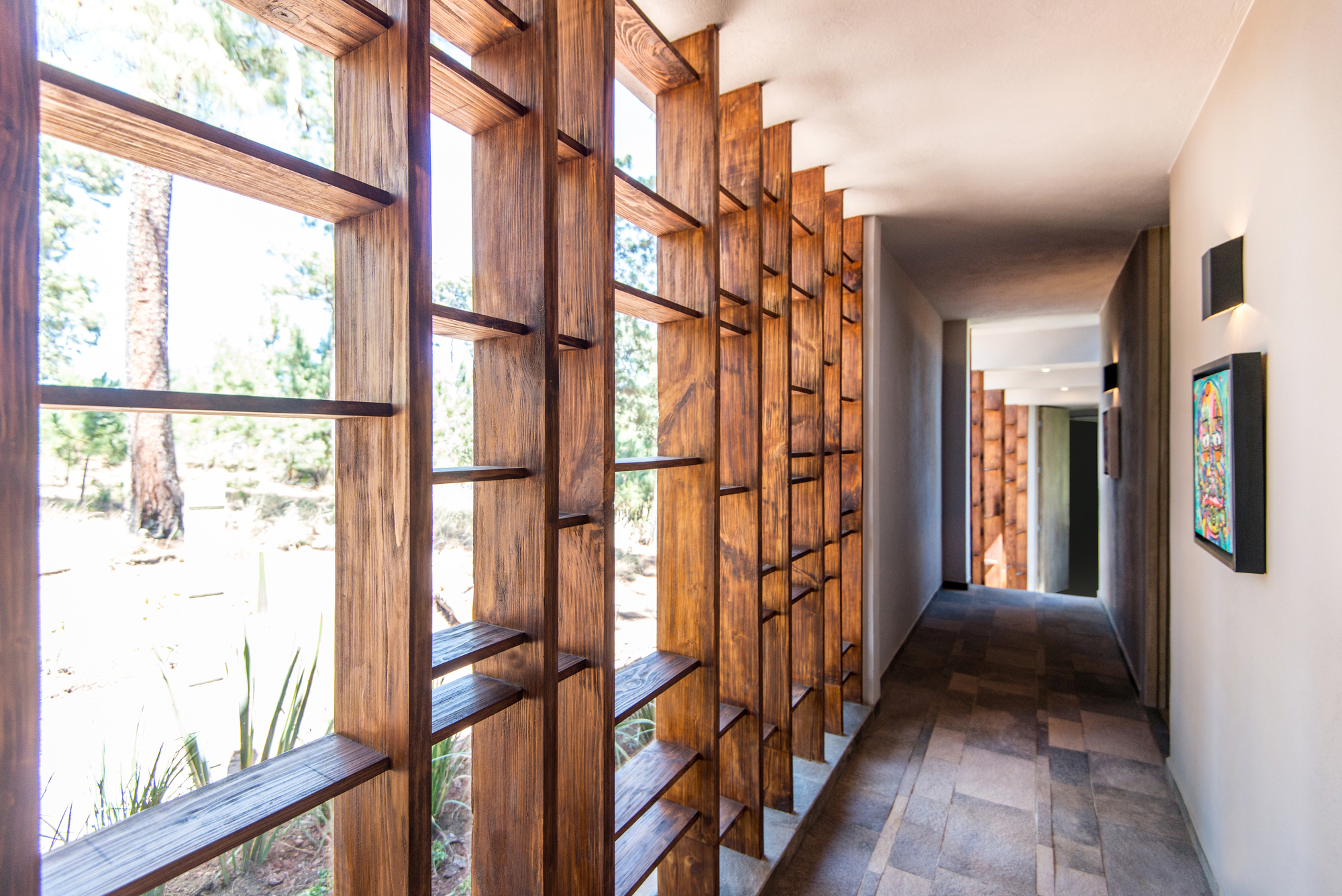 Corridor lined with vertical timber slats framing pine trees outside, with a colorful painting on the far wall in daylight