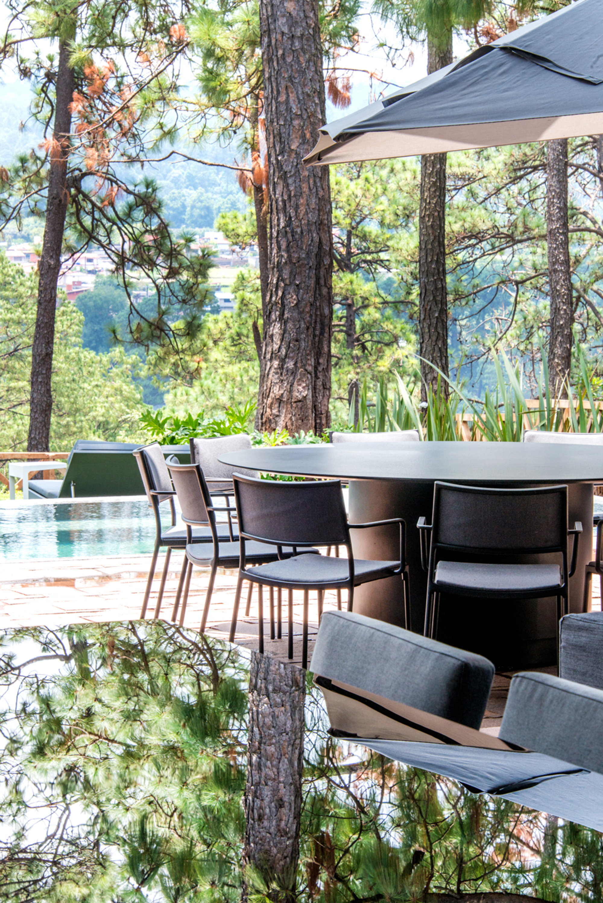 Outdoor dining table with black chairs and umbrella near an infinity pool, pine trunks reflected in the glossy tabletop