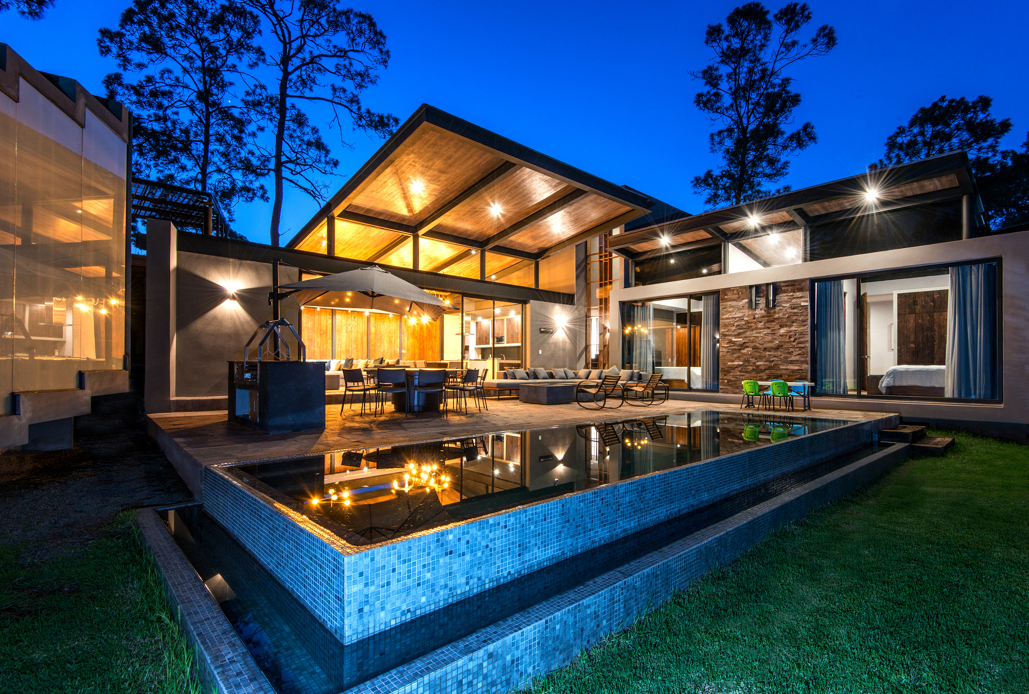Dusk view of the infinity pool with blue mosaic tile edge in front of the glowing timber-roofed terrace and pines