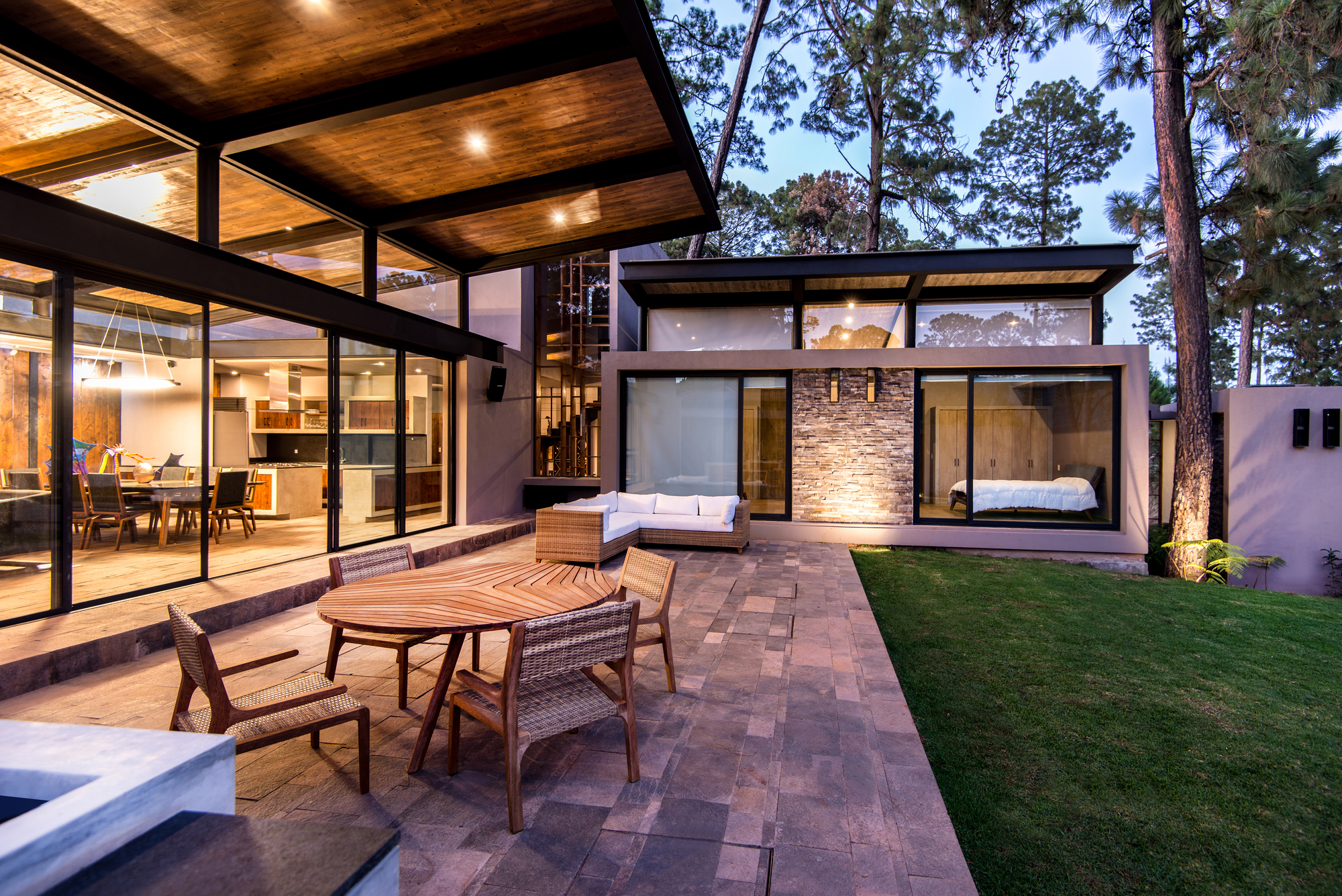 Paved patio with a round teak table and wicker chairs beside the glazed pavilion and stone-clad wall at twilight