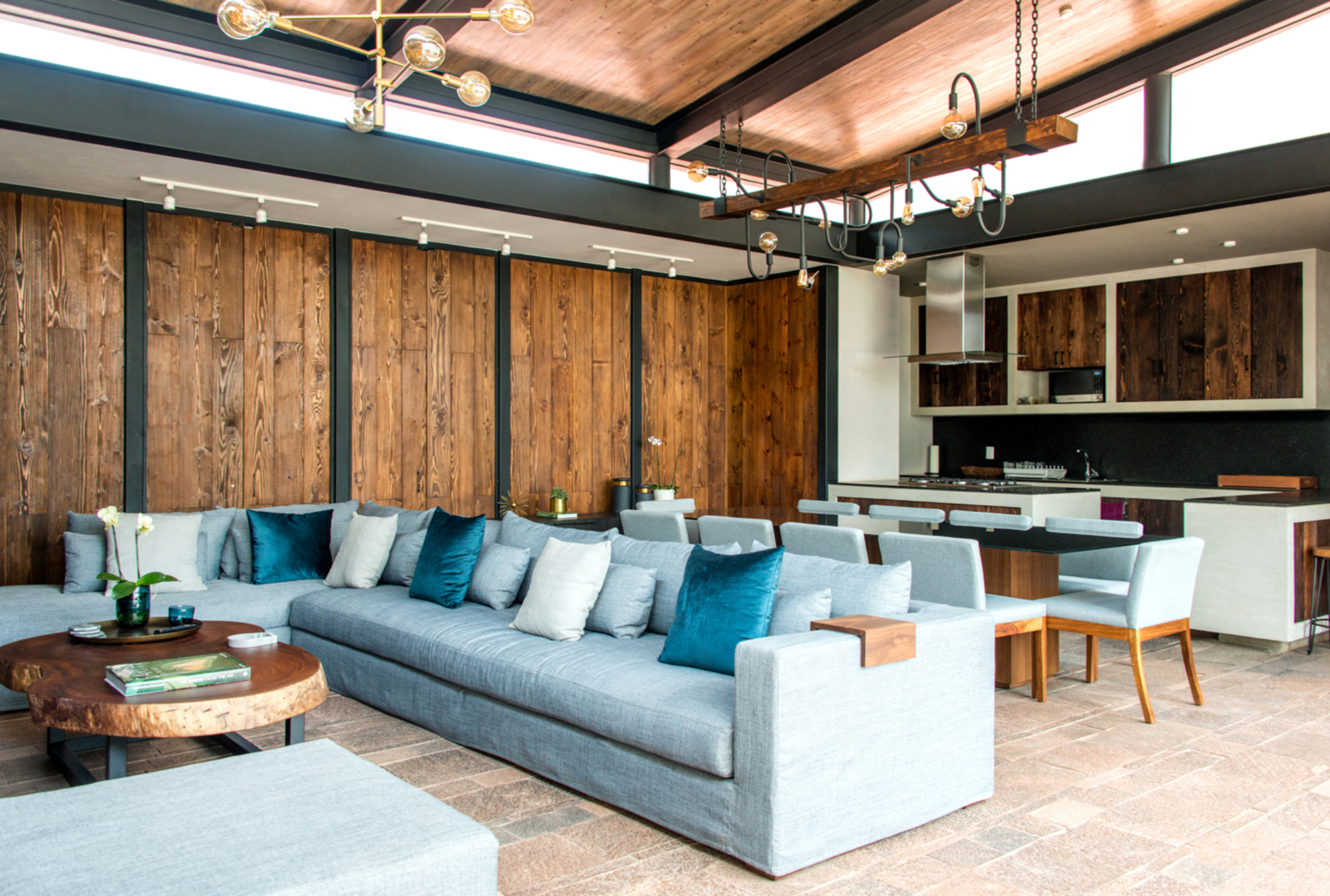 Living area with a grey sofa and teal cushions against reclaimed timber wall panels, with the kitchen beyond
