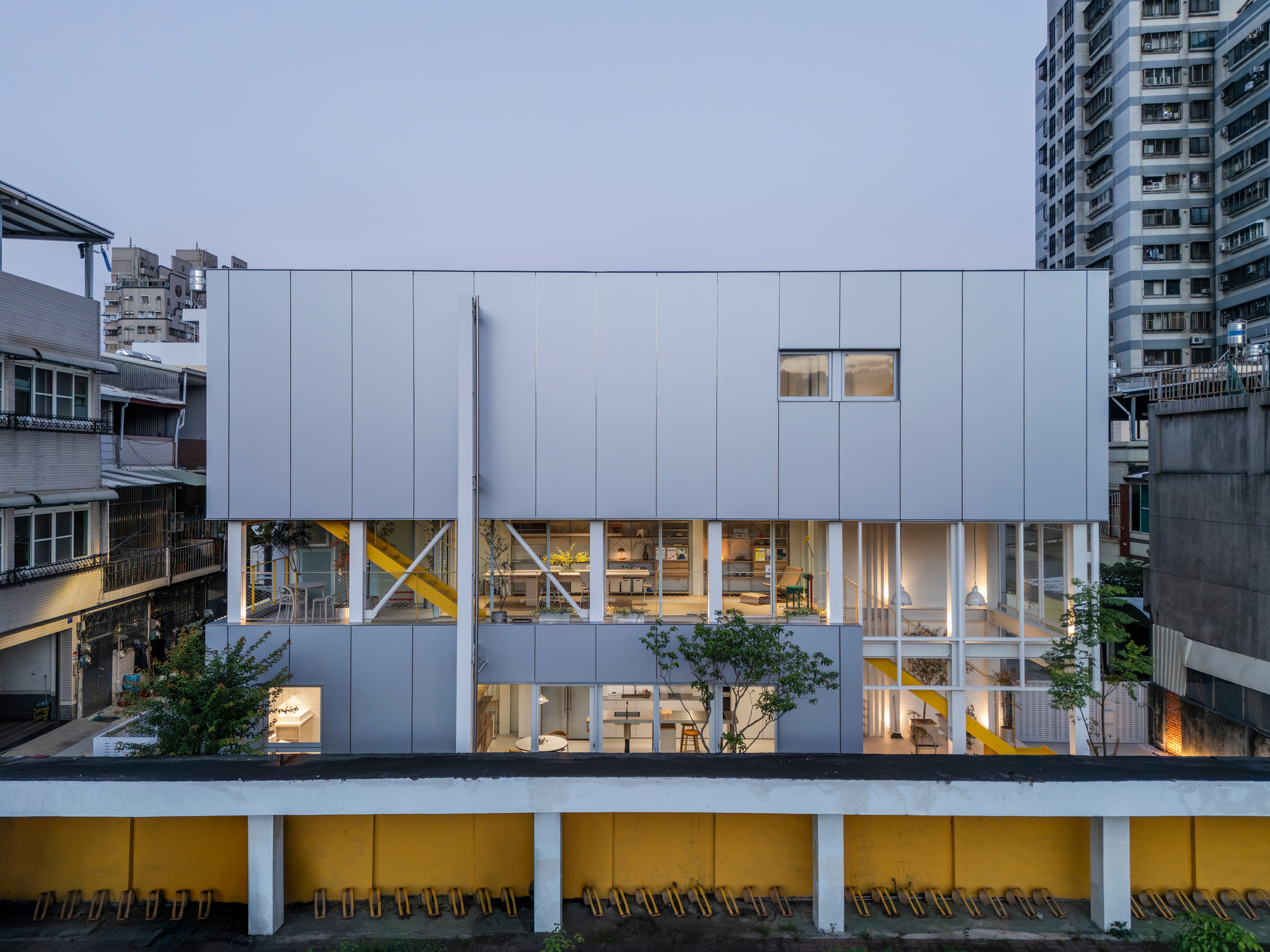 Glazed lower floors and grey panel upper volume seen over a yellow school wall at dusk, with lit interiors and a yellow stair