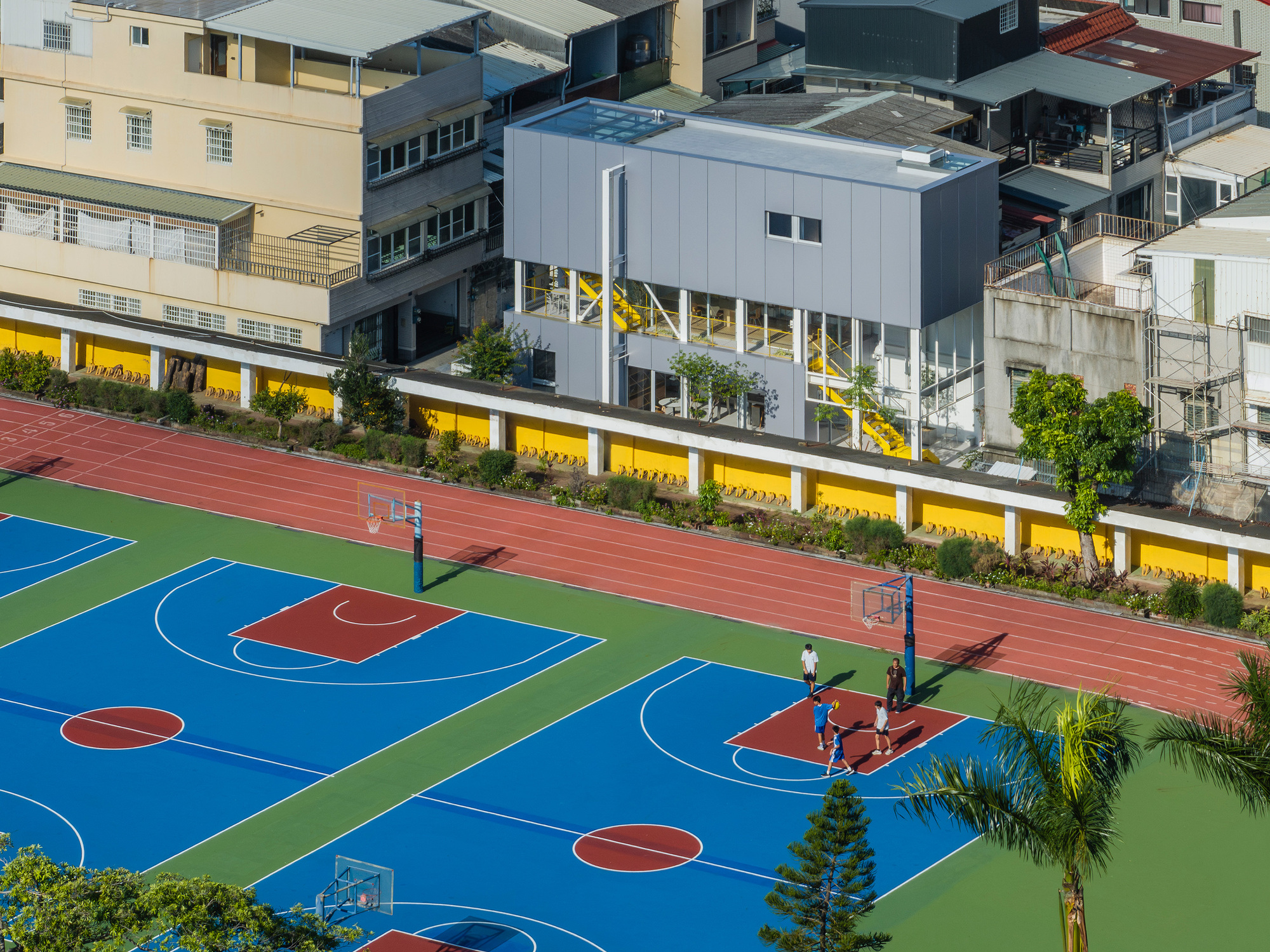 Aerial view of the grey paneled building behind a yellow wall and running track, with players on blue basketball courts in sunlight