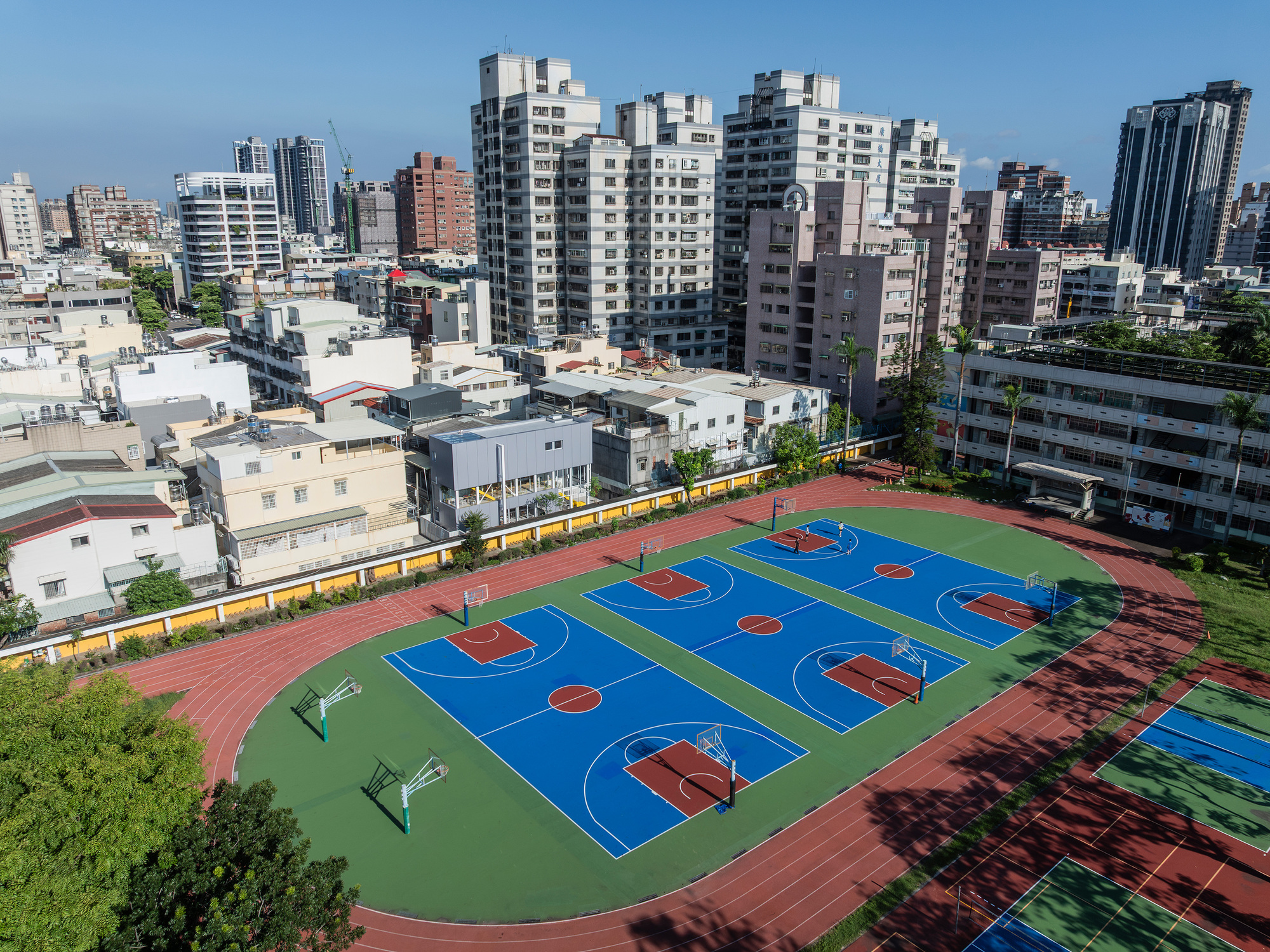 Aerial view of blue basketball courts inside a red running track, surrounded by apartment towers under a clear blue sky