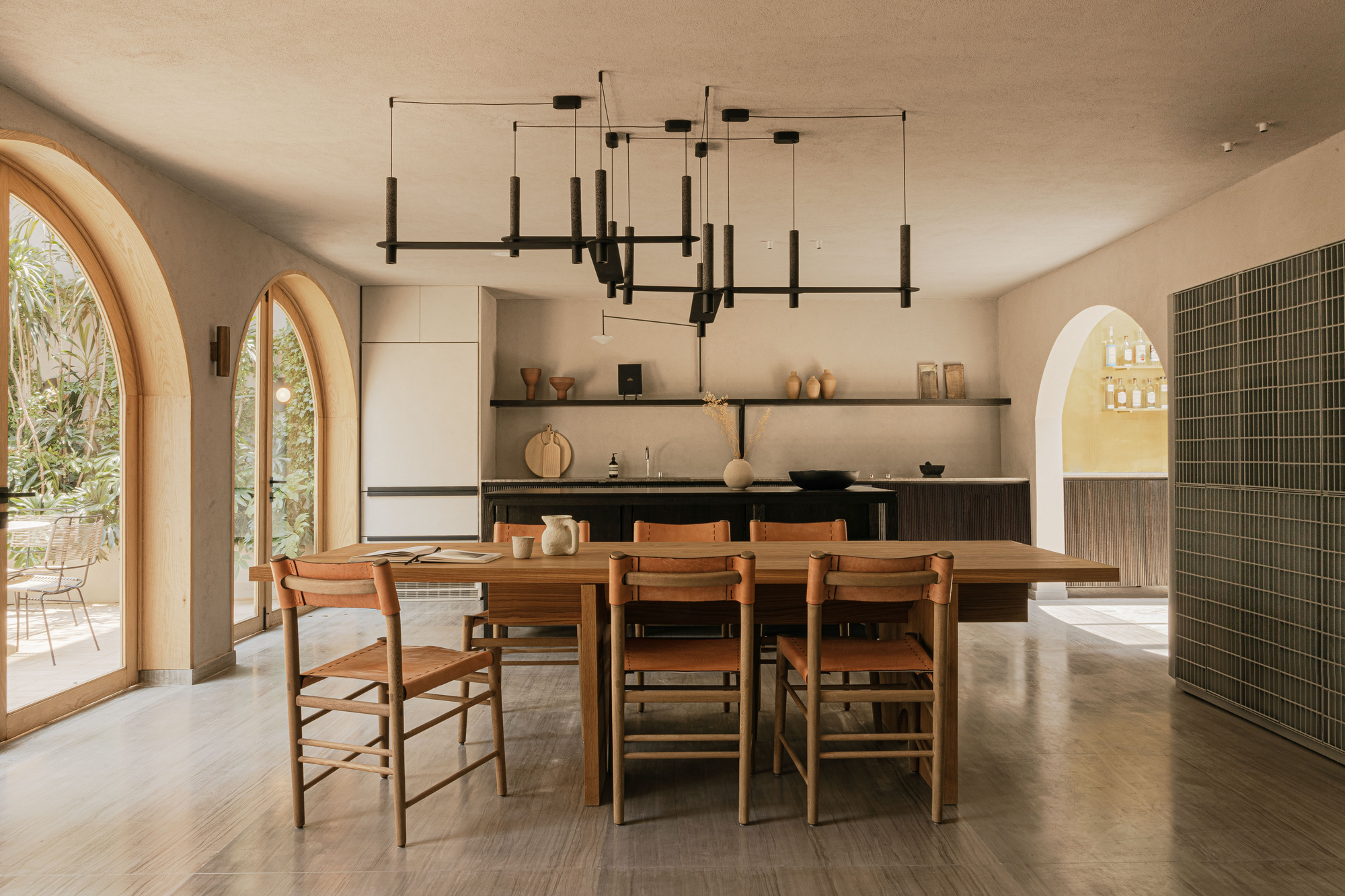 Open dining room with a long timber table, leather-seat chairs and arched glazed doors opening to a sunlit garden