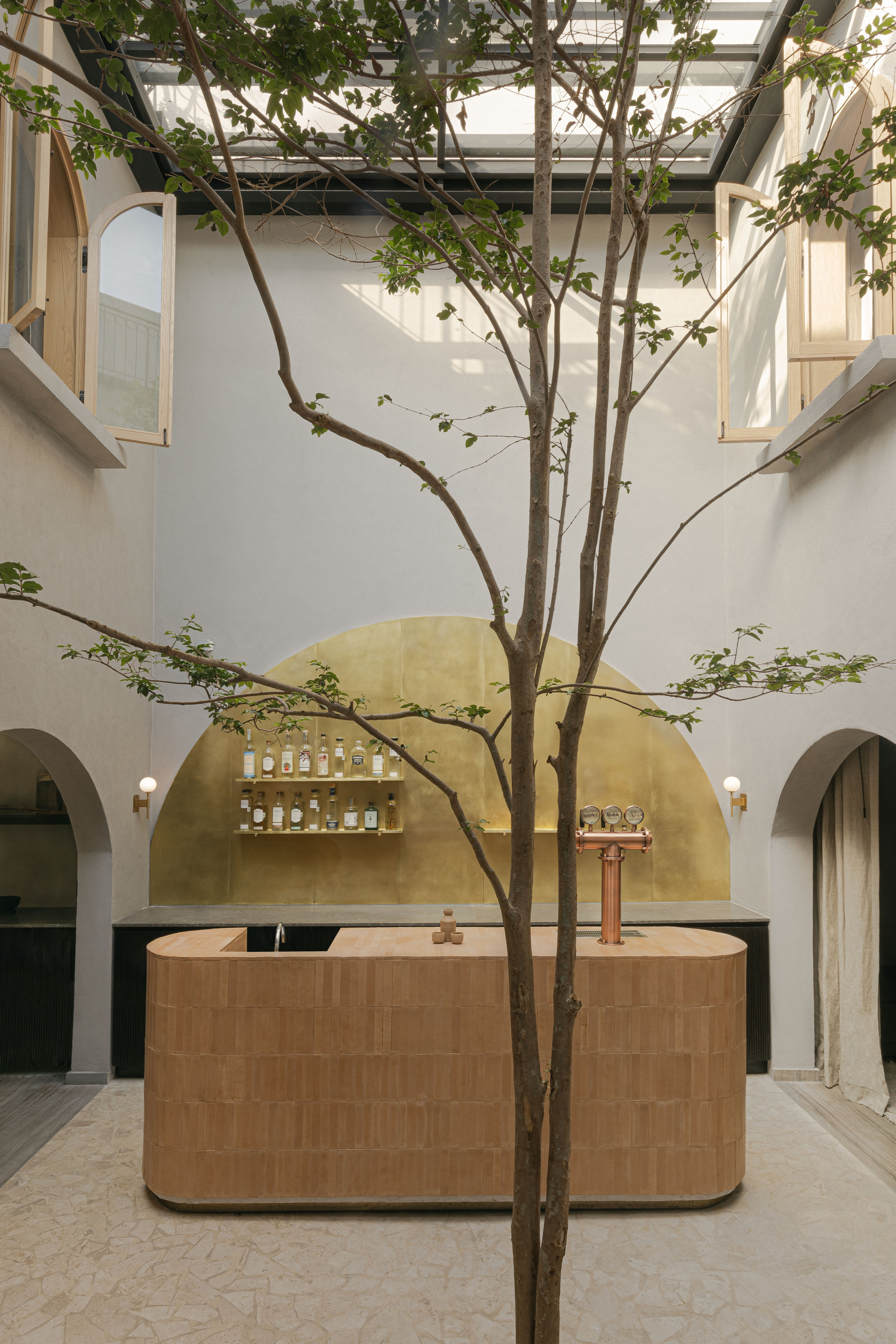Rounded brick-clad bar and brass arched backdrop in a skylit courtyard, with a slender tree in the foreground
