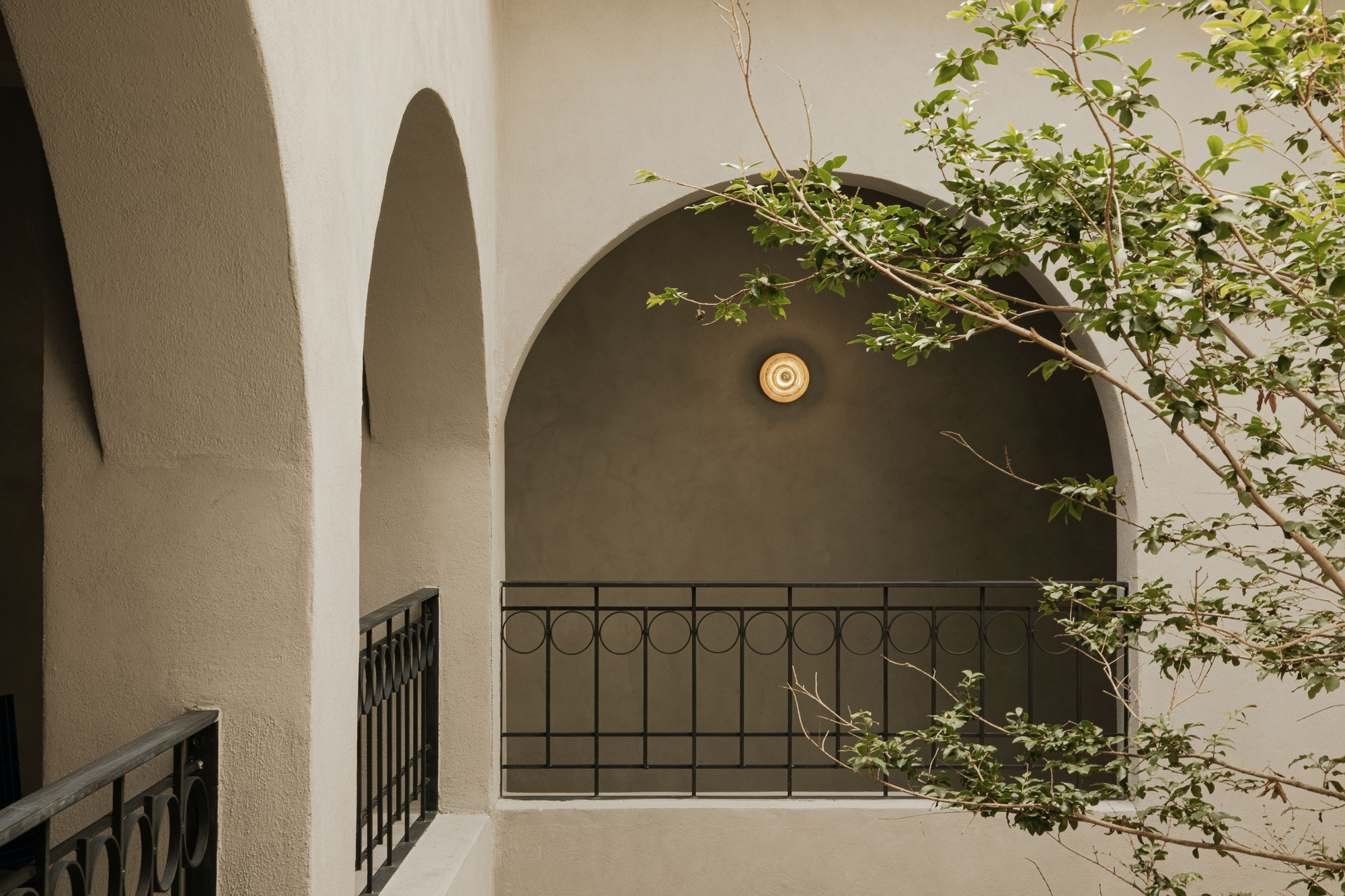 Plastered arched balcony with a black iron railing and round wall lamp, framed by leafy branches in soft light