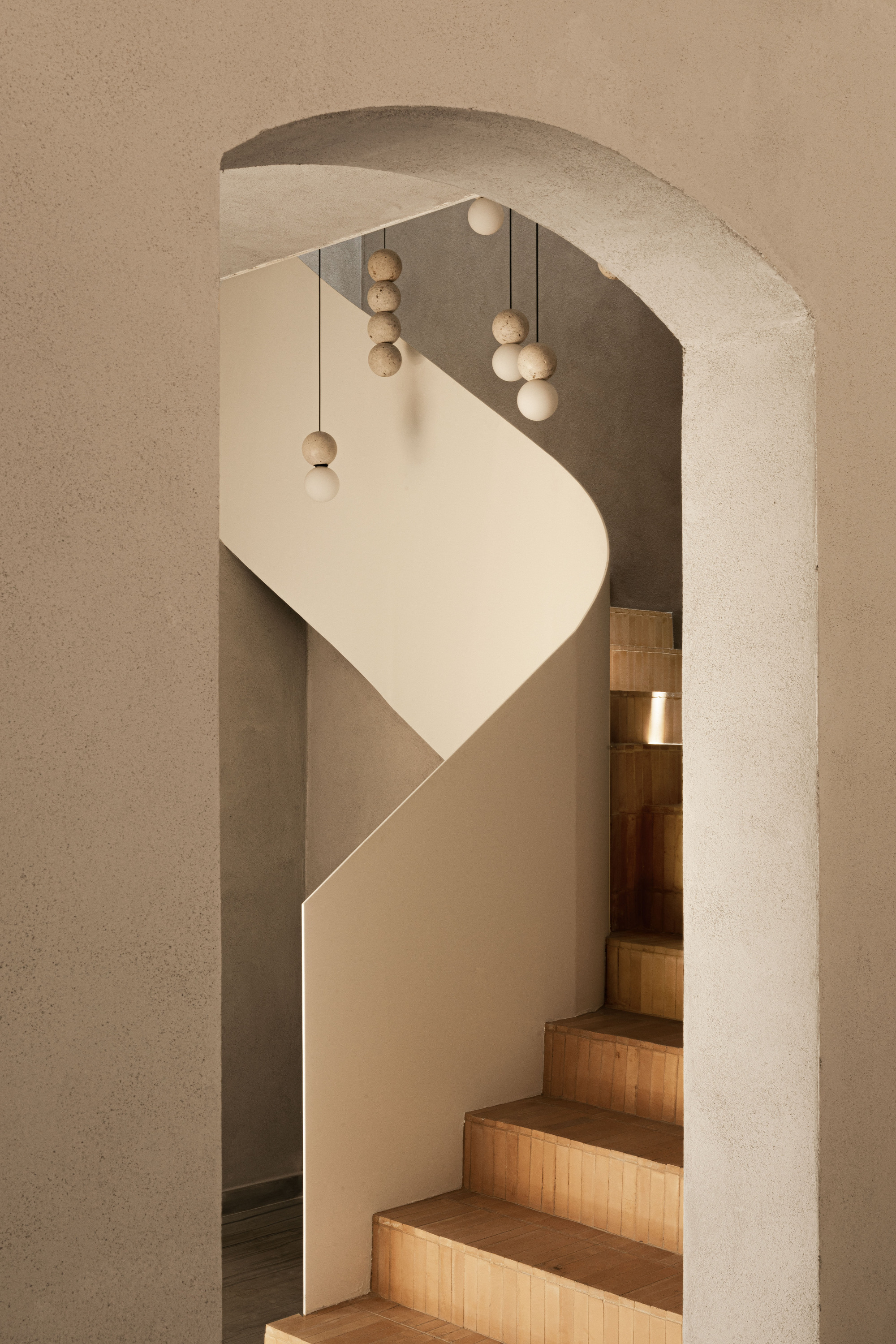 Curving white stair wall seen through a rough plaster arch, with stone bead pendants hanging above timber-clad steps