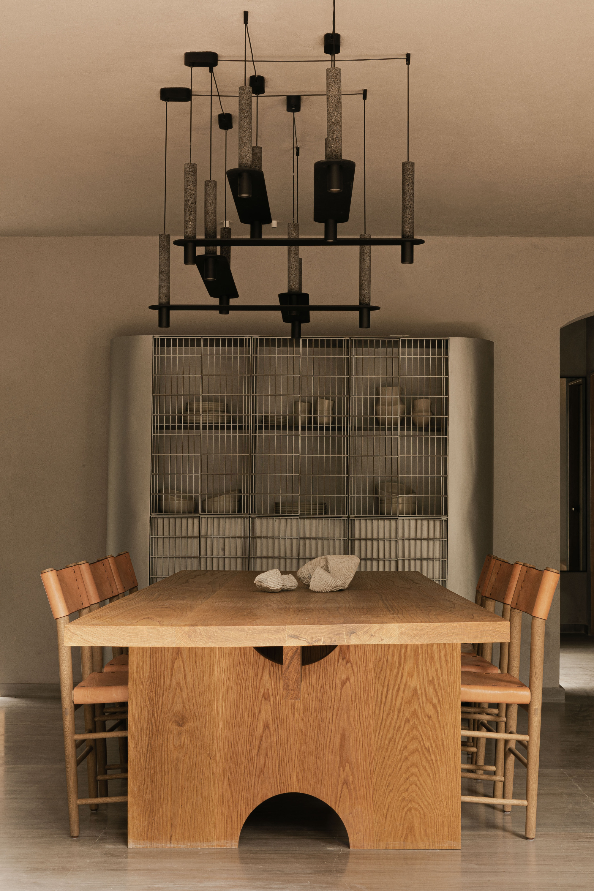 Solid oak dining table with an arched base cutout, woven-back chairs and a black pendant cluster before a mesh-fronted steel cabinet
