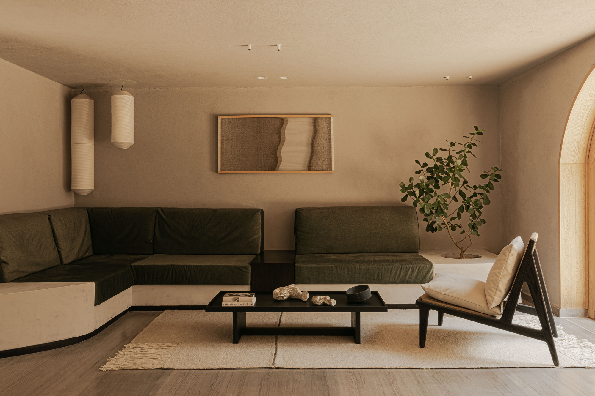 Curved olive-green sectional sofa facing a low black table, with a potted tree and paper lanterns in soft daylight