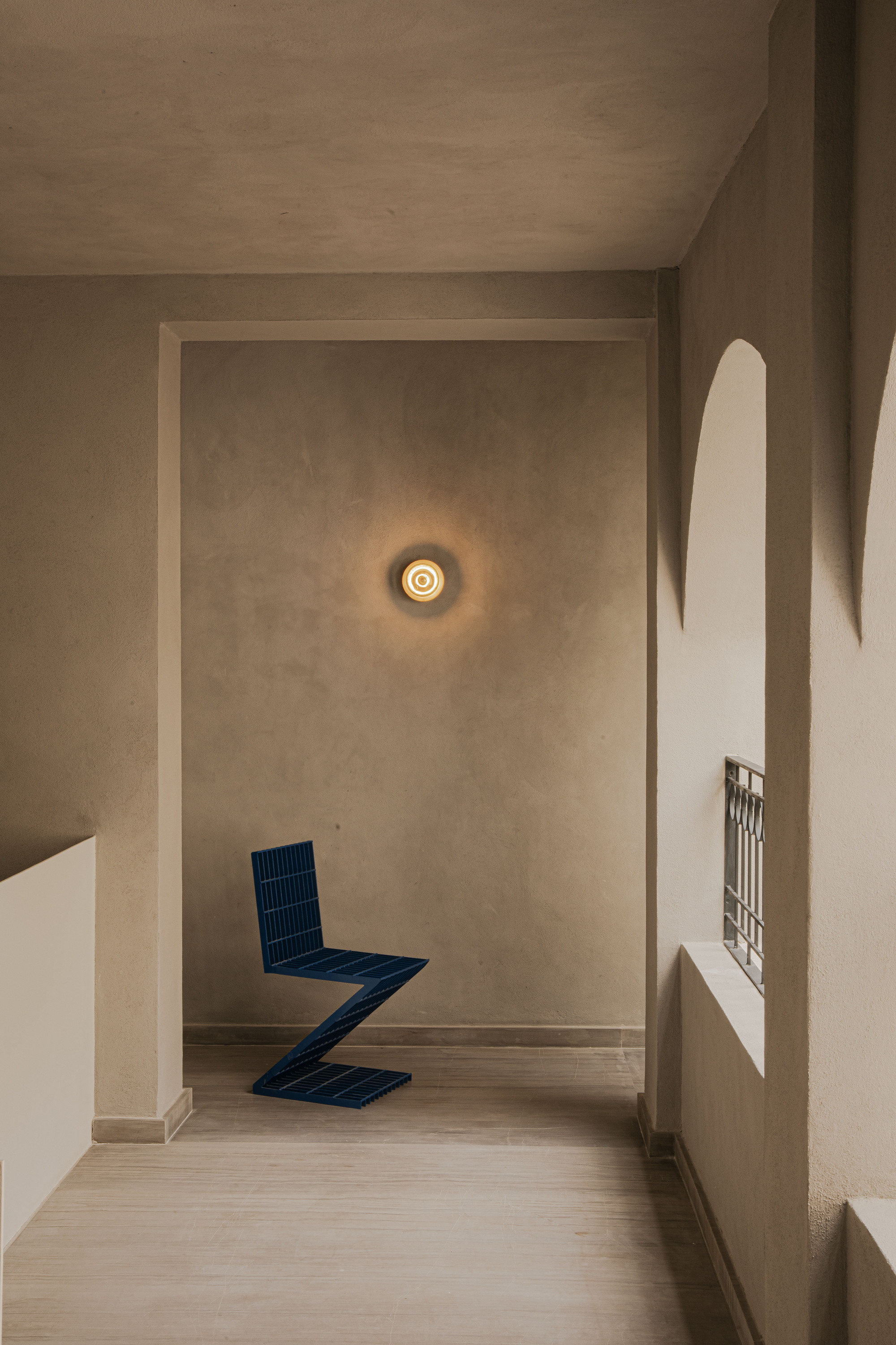 Blue zigzag chair standing in a plastered alcove beneath a round glowing wall light, beside arched openings