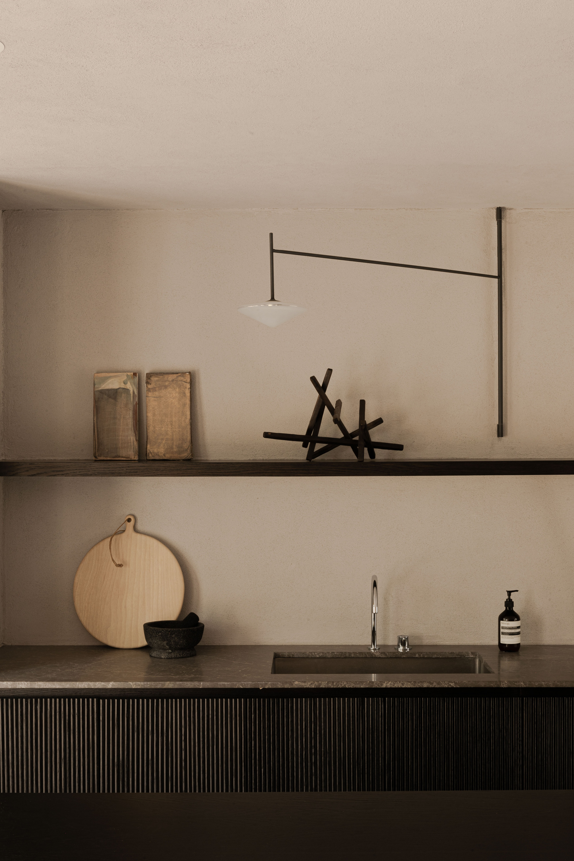 Kitchen counter with fluted dark front, steel sink, round wooden board and a long-armed wall lamp over a shelf