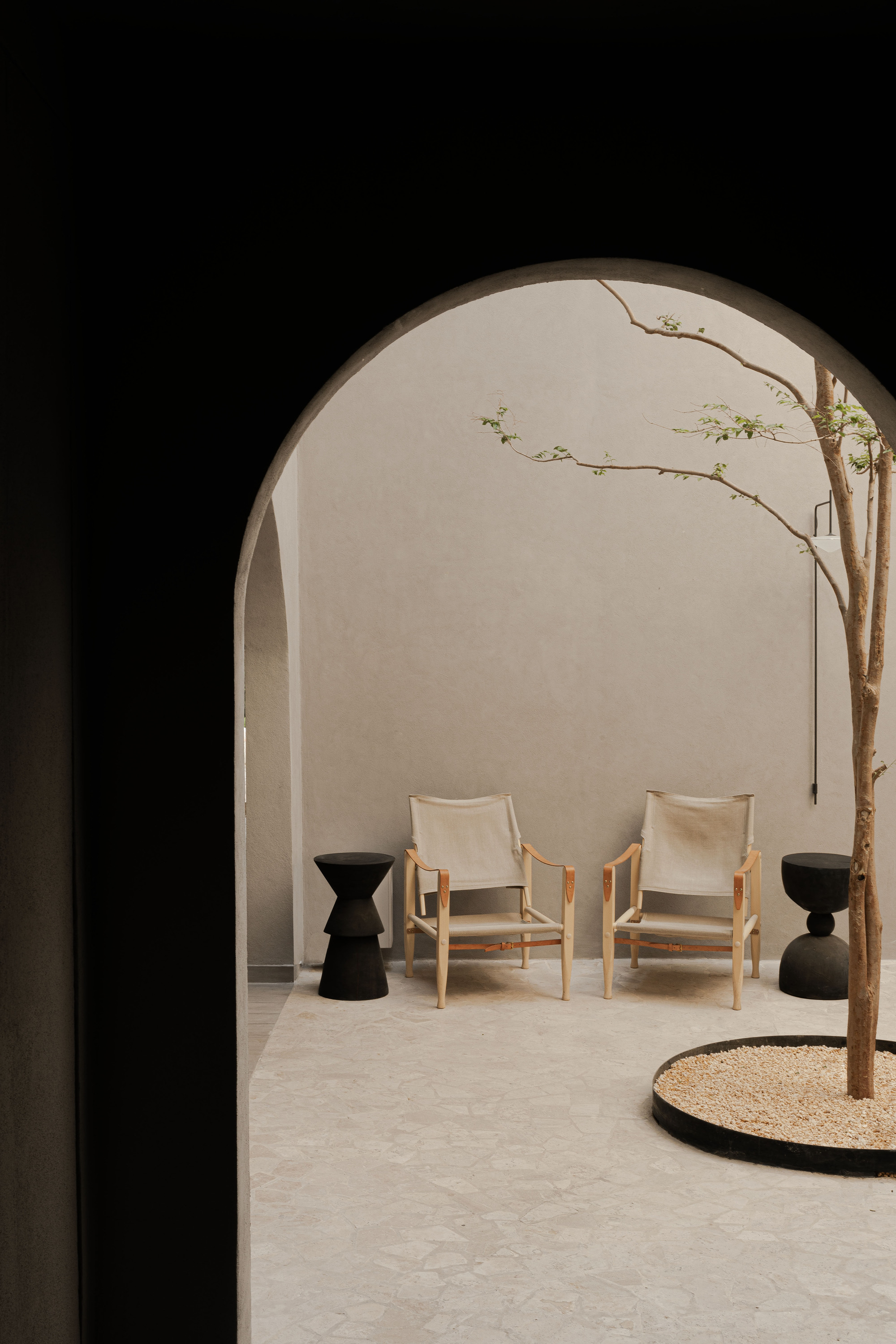 View through a dark arch into a courtyard with two canvas chairs, black stools and a slender tree in a gravel bed
