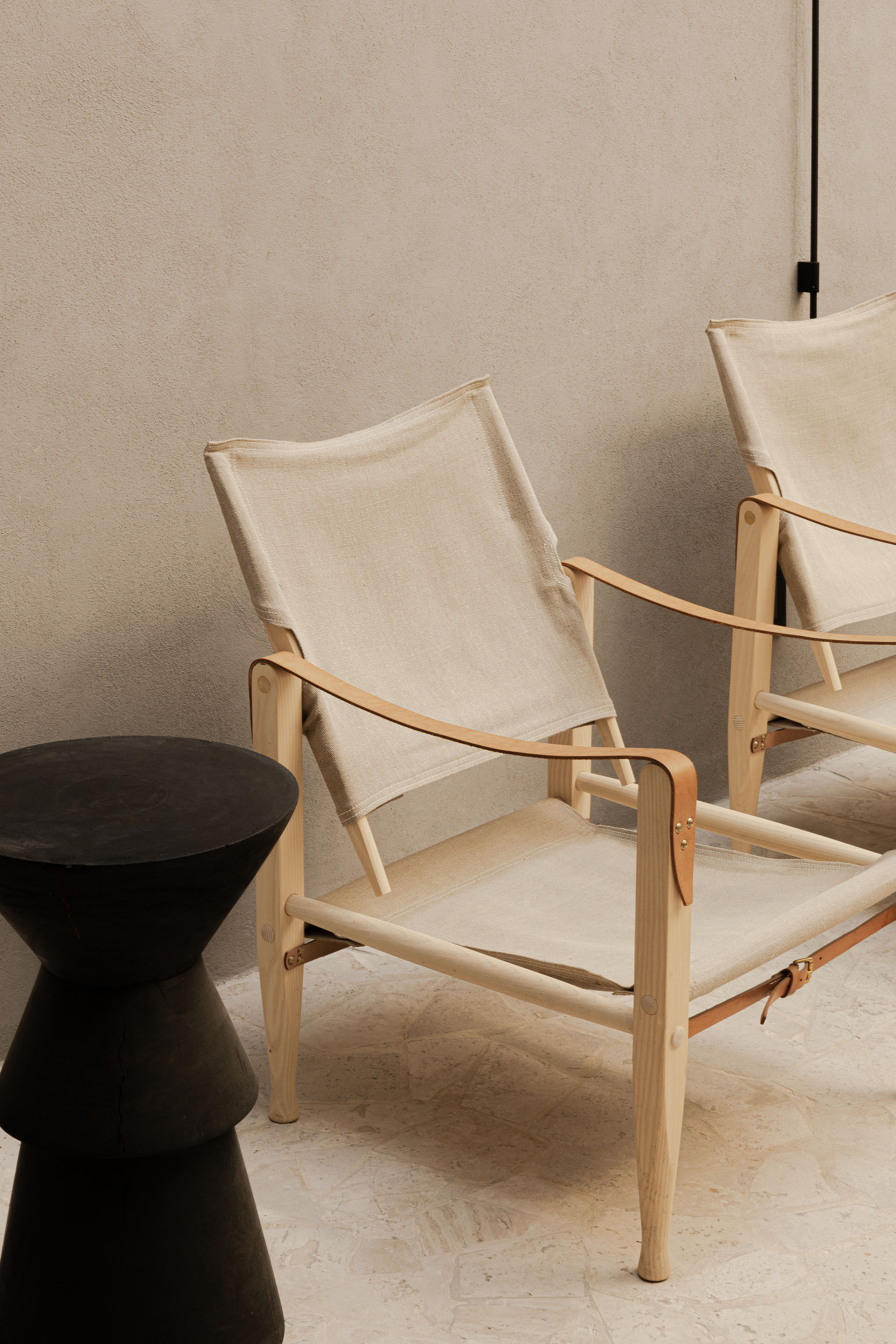 Two canvas and ash safari chairs with leather straps beside a black sculptural side table on a pale stone floor