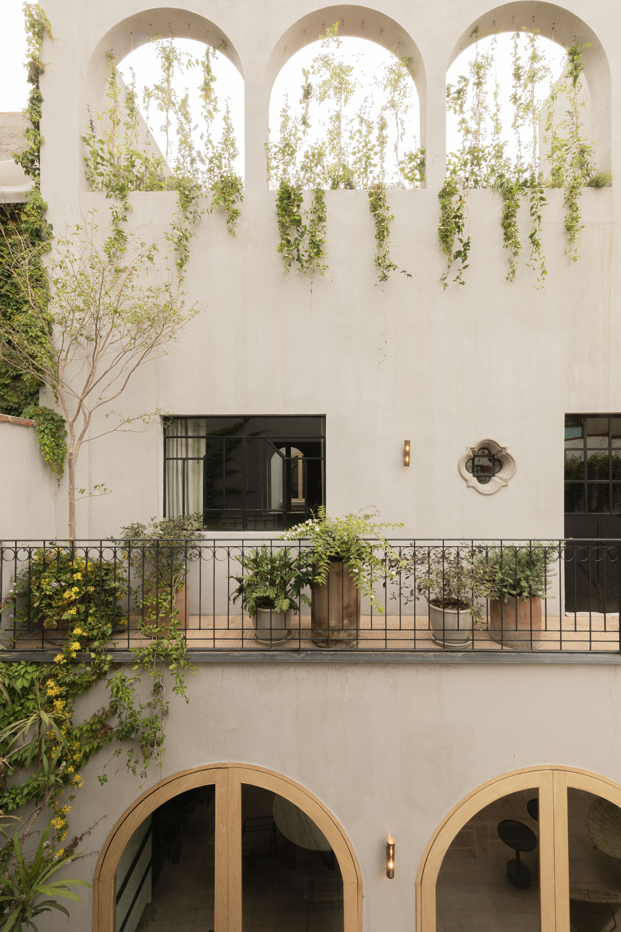 Plastered courtyard facade with arched openings, a wrought iron balcony of potted plants and vines trailing down in soft daylight
