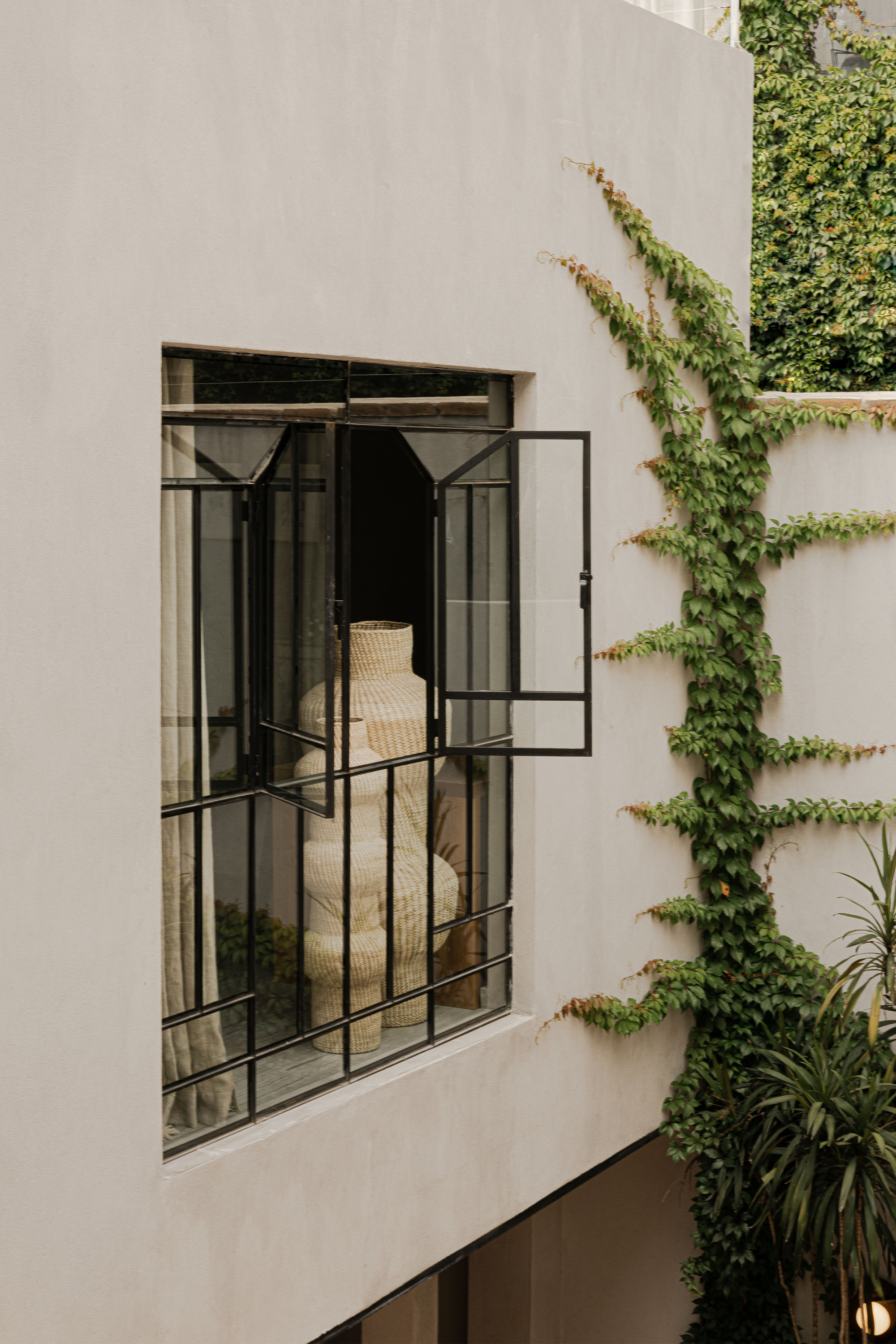 Open steel-framed window in a plaster wall showing a woven vase inside, with ivy climbing the adjacent wall