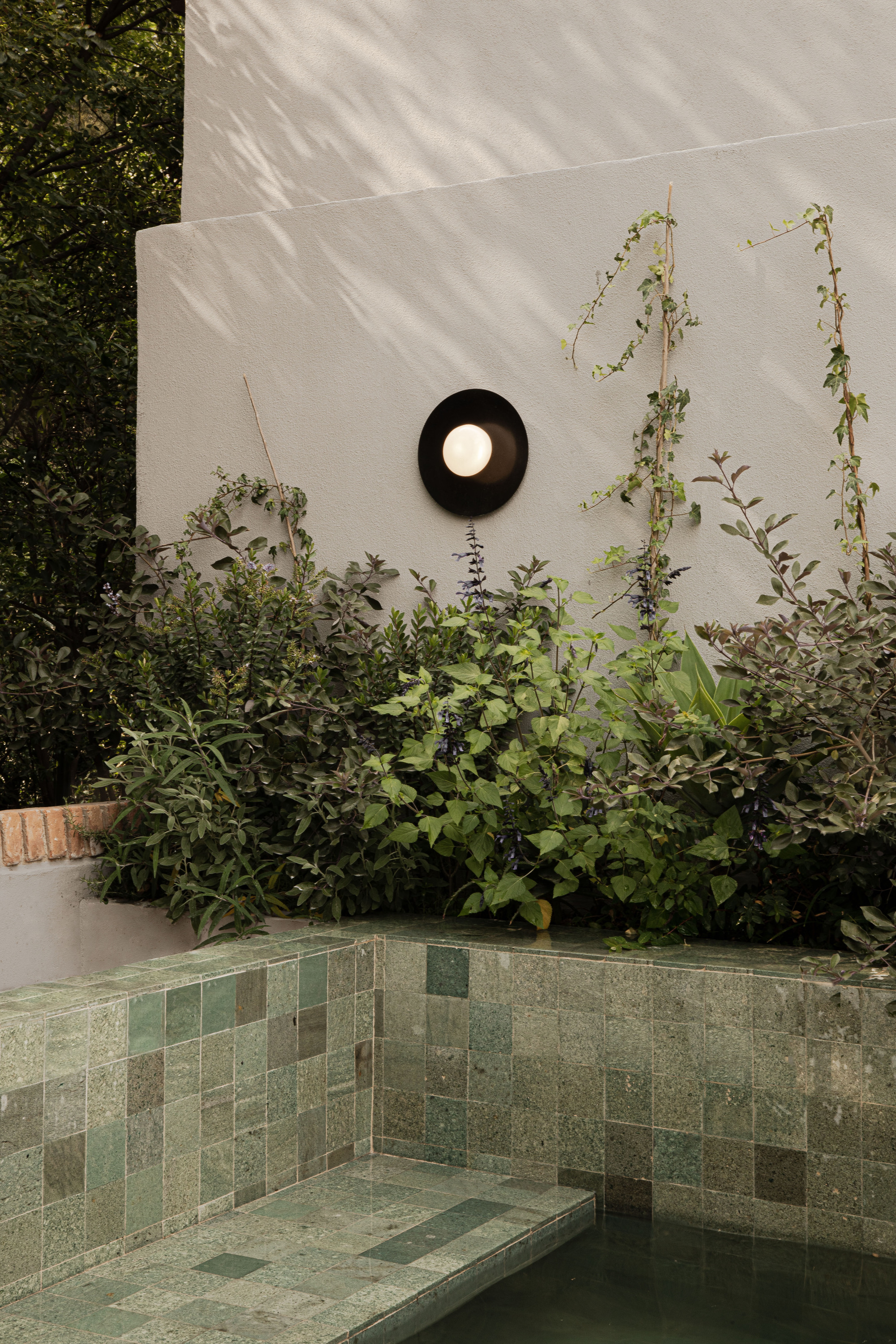 Green glazed tile plunge pool edge beneath a round black wall lamp, with dappled sunlight and leafy planting behind