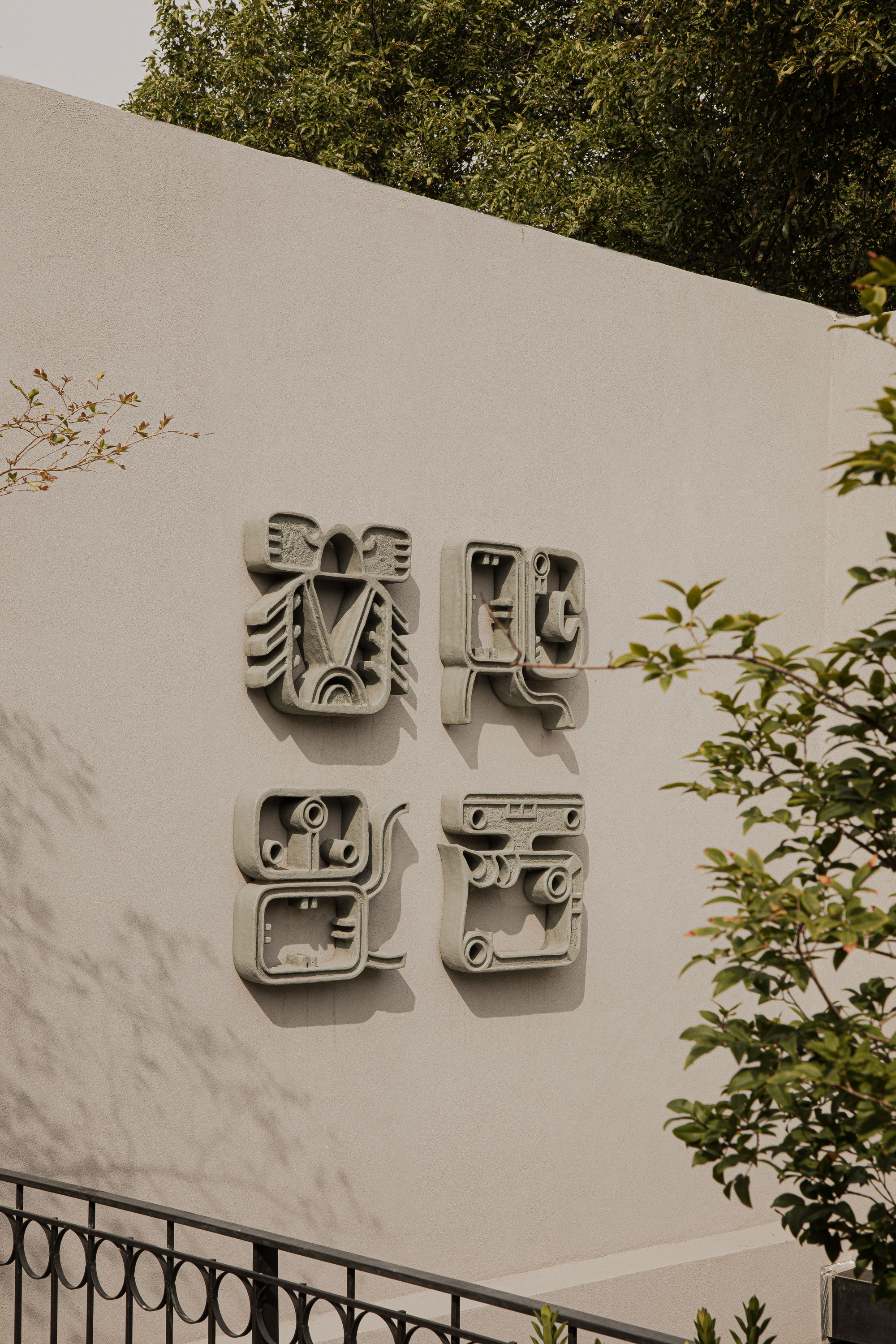 Four carved stone glyph-like reliefs mounted on a plaster wall above an iron railing, with tree branches in the foreground