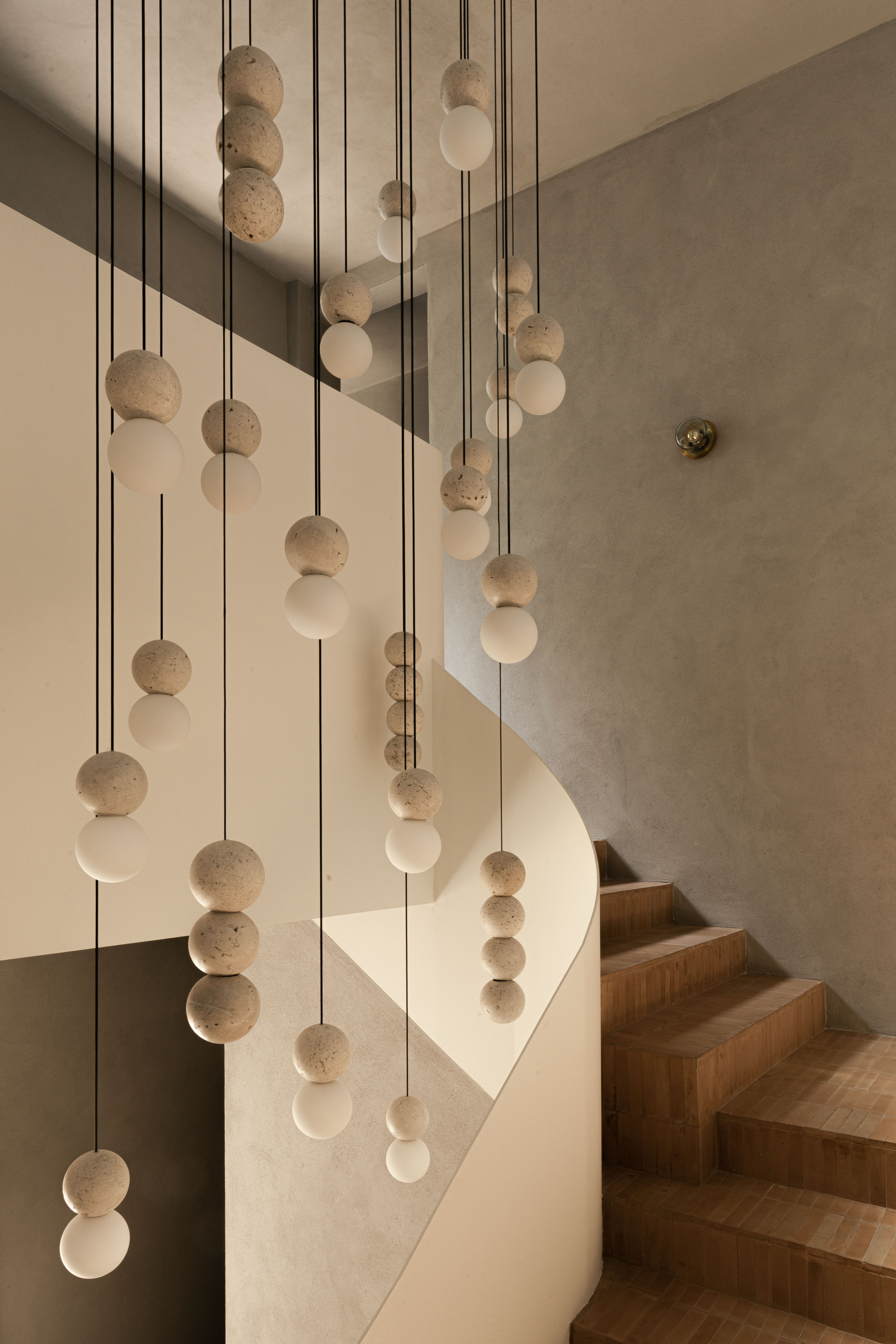 Cluster of travertine and white glass spherical pendants hanging beside a curved plaster stair wall with terracotta-tiled steps in warm light