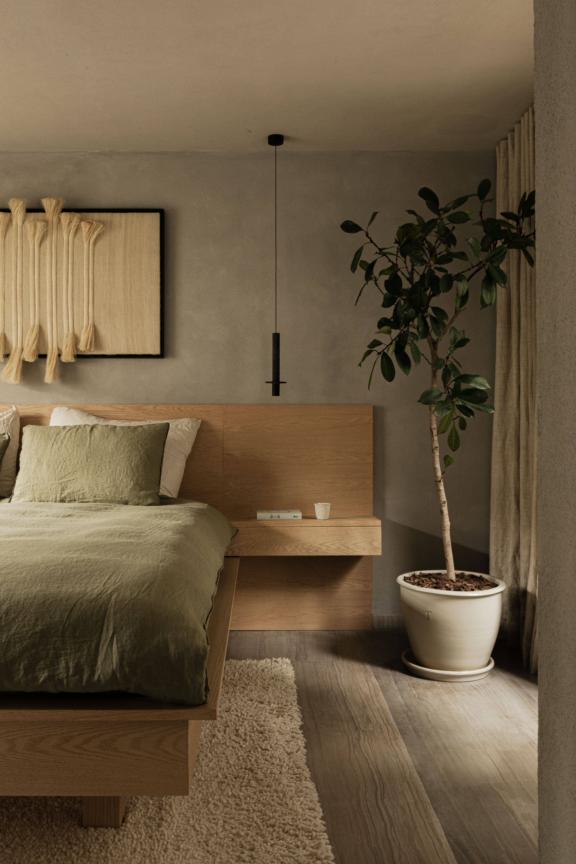 Oak platform bed with floating bedside shelf, green linen and a potted ficus beside a linen curtain in soft daylight