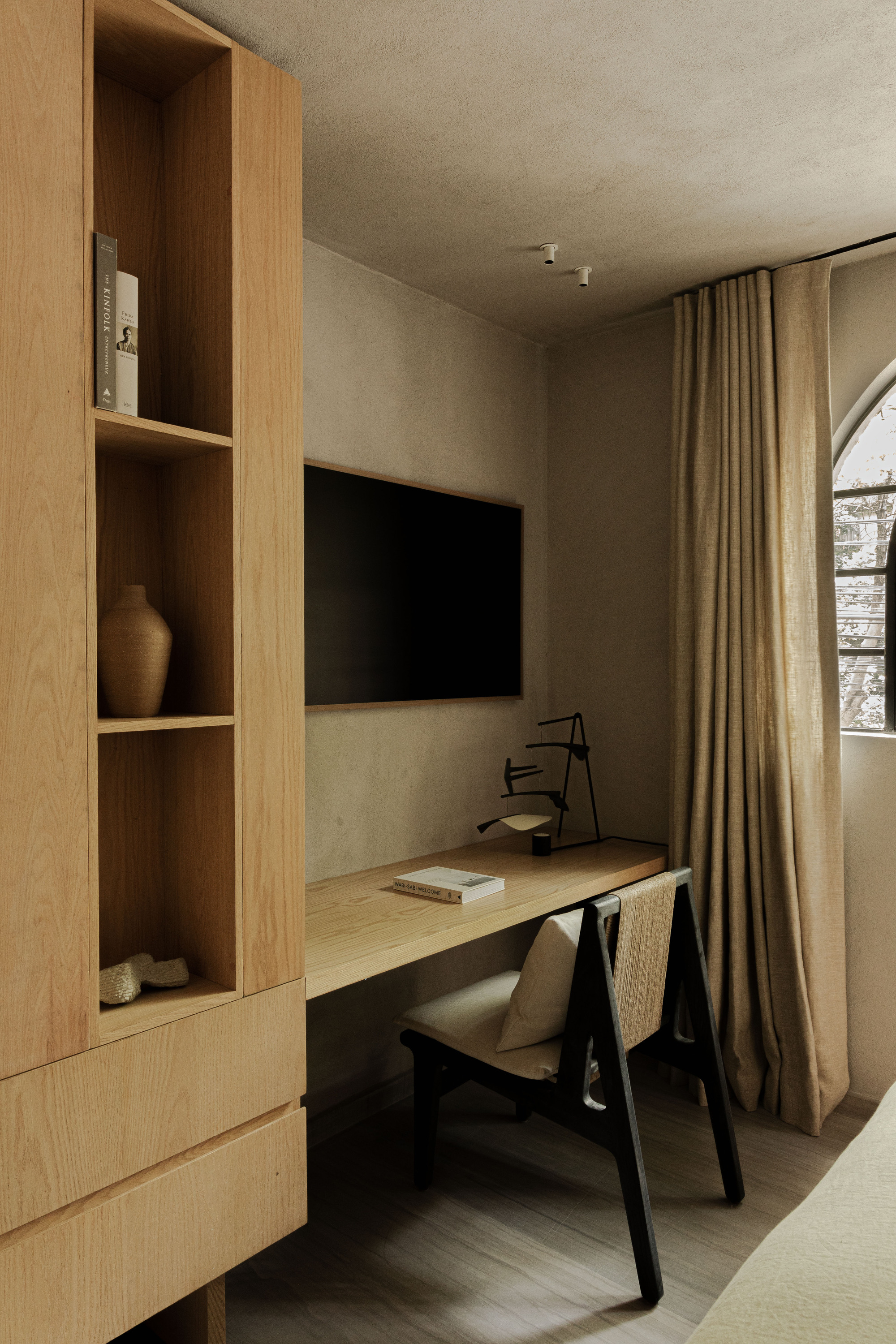 Oak shelving unit and built-in desk with a black chair beneath an arched window and a tall linen curtain