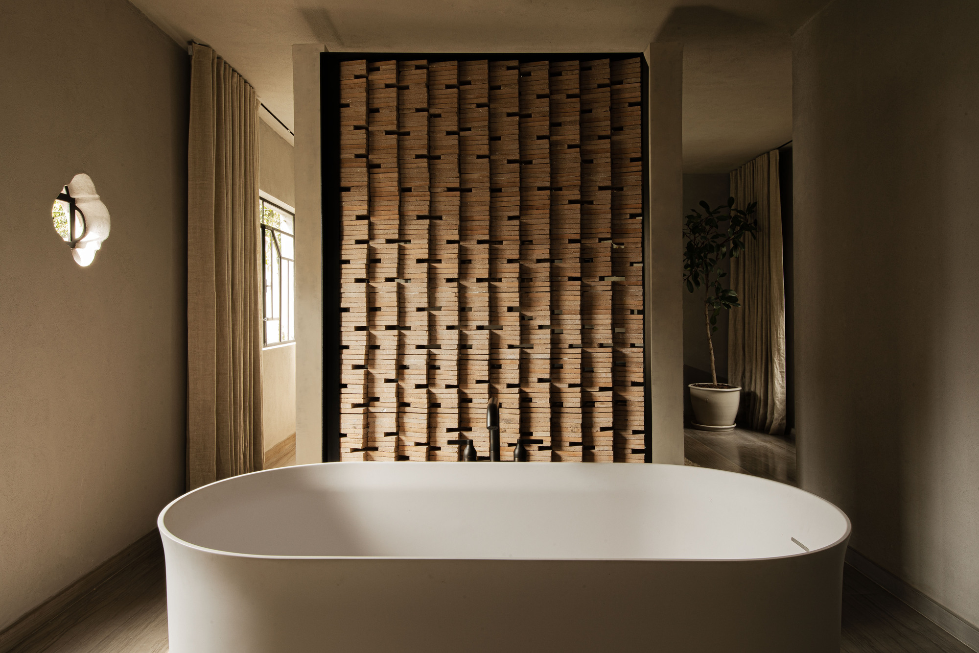 Freestanding white tub in front of a stacked terracotta tile screen, with a potted tree reflected in a mirror at right