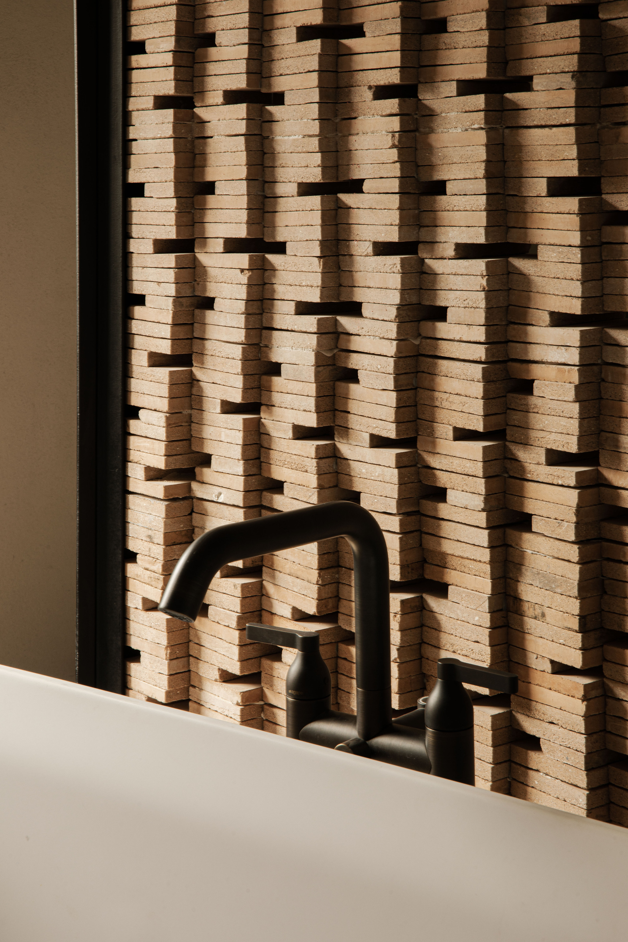 Close-up of a black bath faucet against a stacked terracotta tile screen with dark gaps, lit by low side light