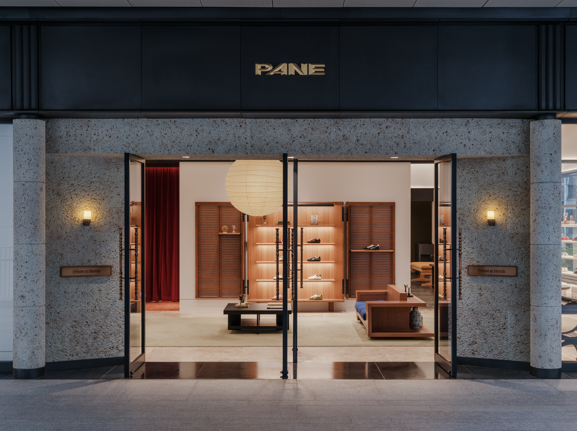 Shopfront entrance with textured stone cladding, open black-framed glass doors and wall sconces glowing at dusk