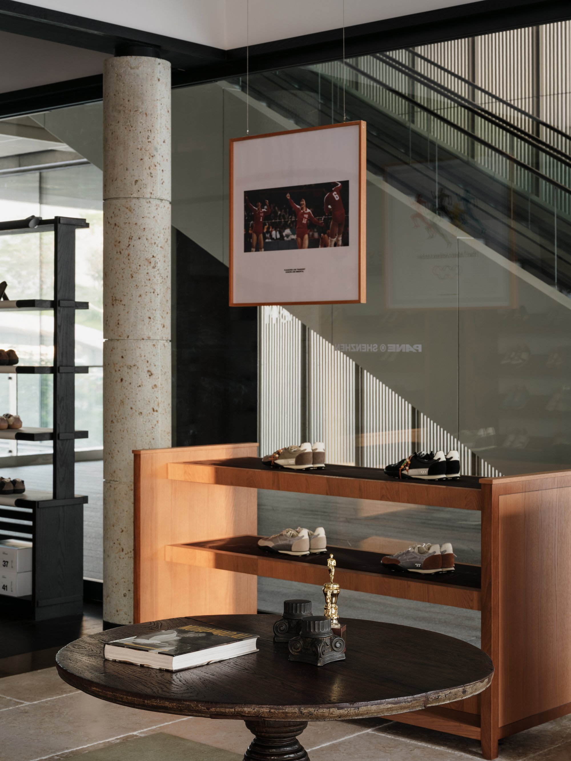 Round dark wood table with a book and trophy beside a stone column and timber shelf, with a glass-backed staircase behind