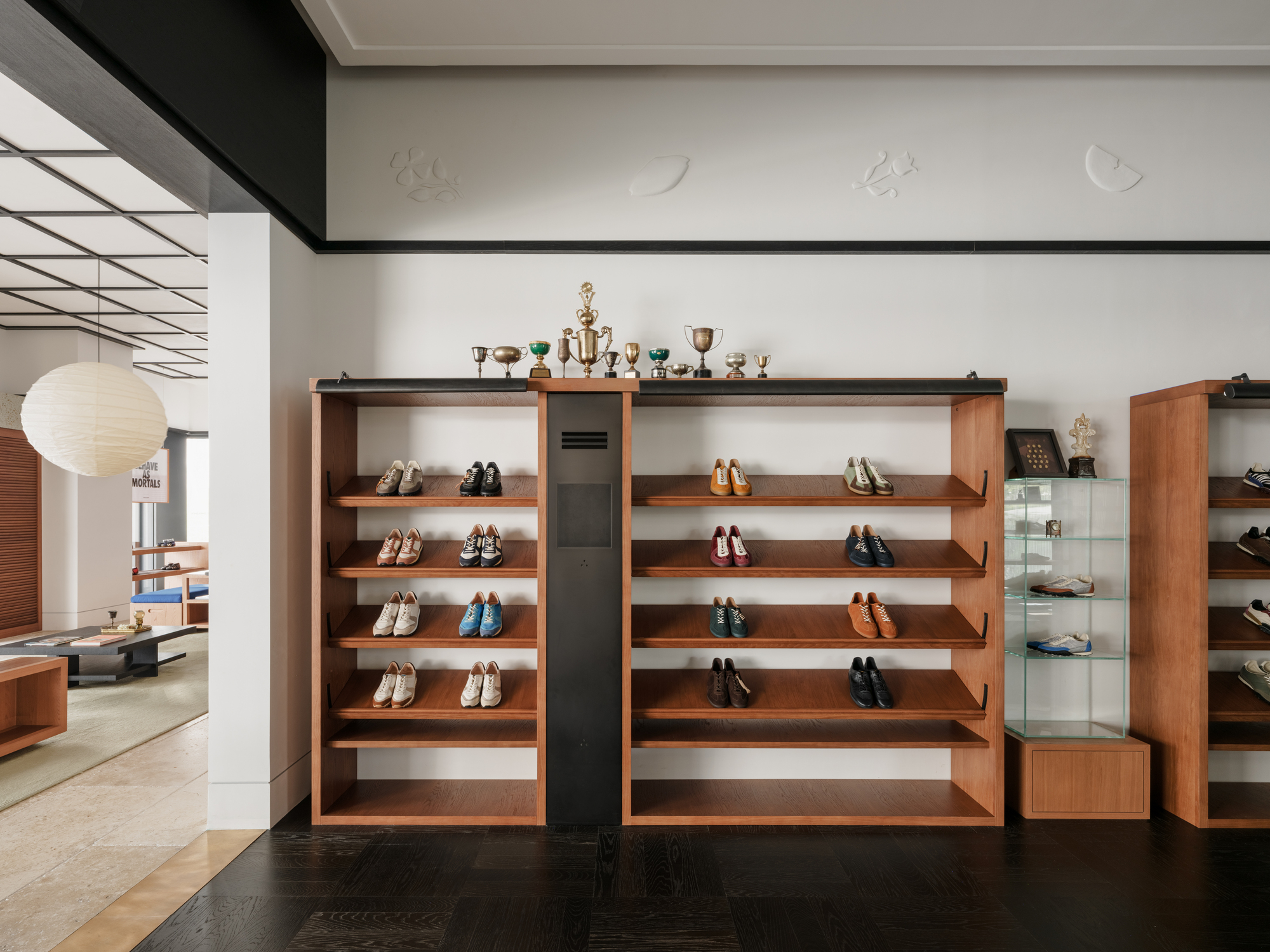 Timber shoe shelving unit topped with small trophies against a white wall with relief carvings, next to a glass display case