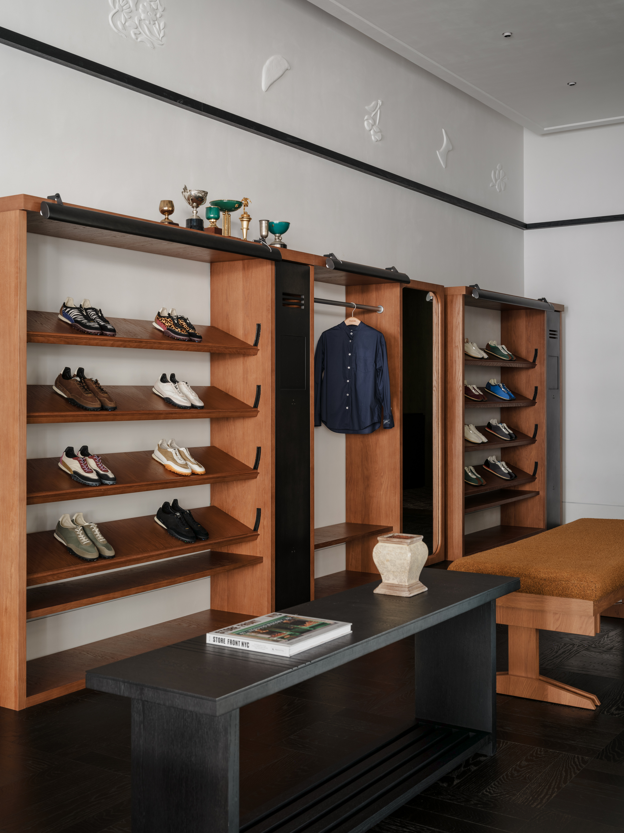 Timber shelving with sneakers, a hanging navy shirt and a mirror panel, with a dark bench and an ochre upholstered seat in front