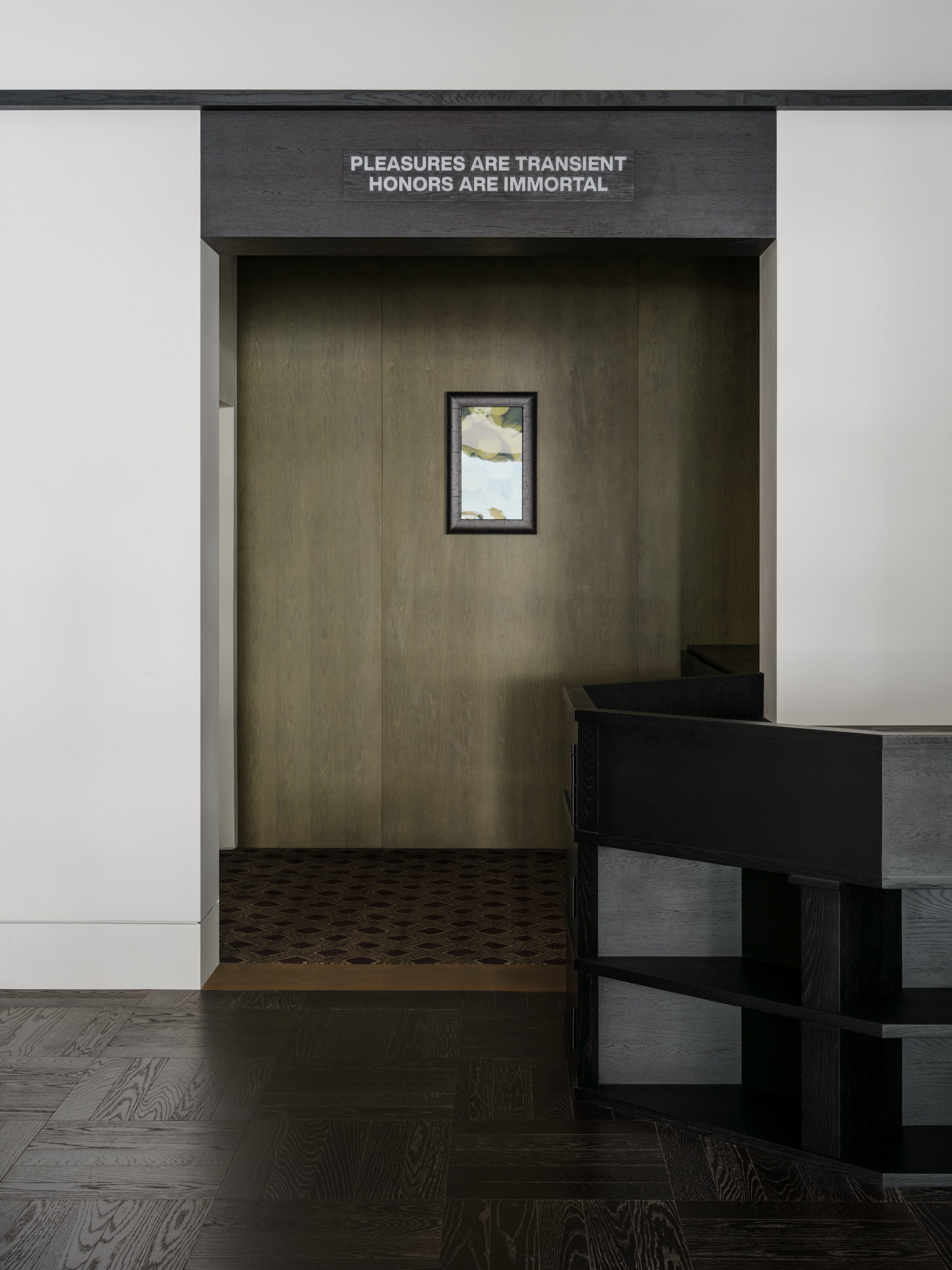 Dark framed doorway with lettered sign above an olive wood-paneled alcove holding a small framed picture, beside a black shelving unit