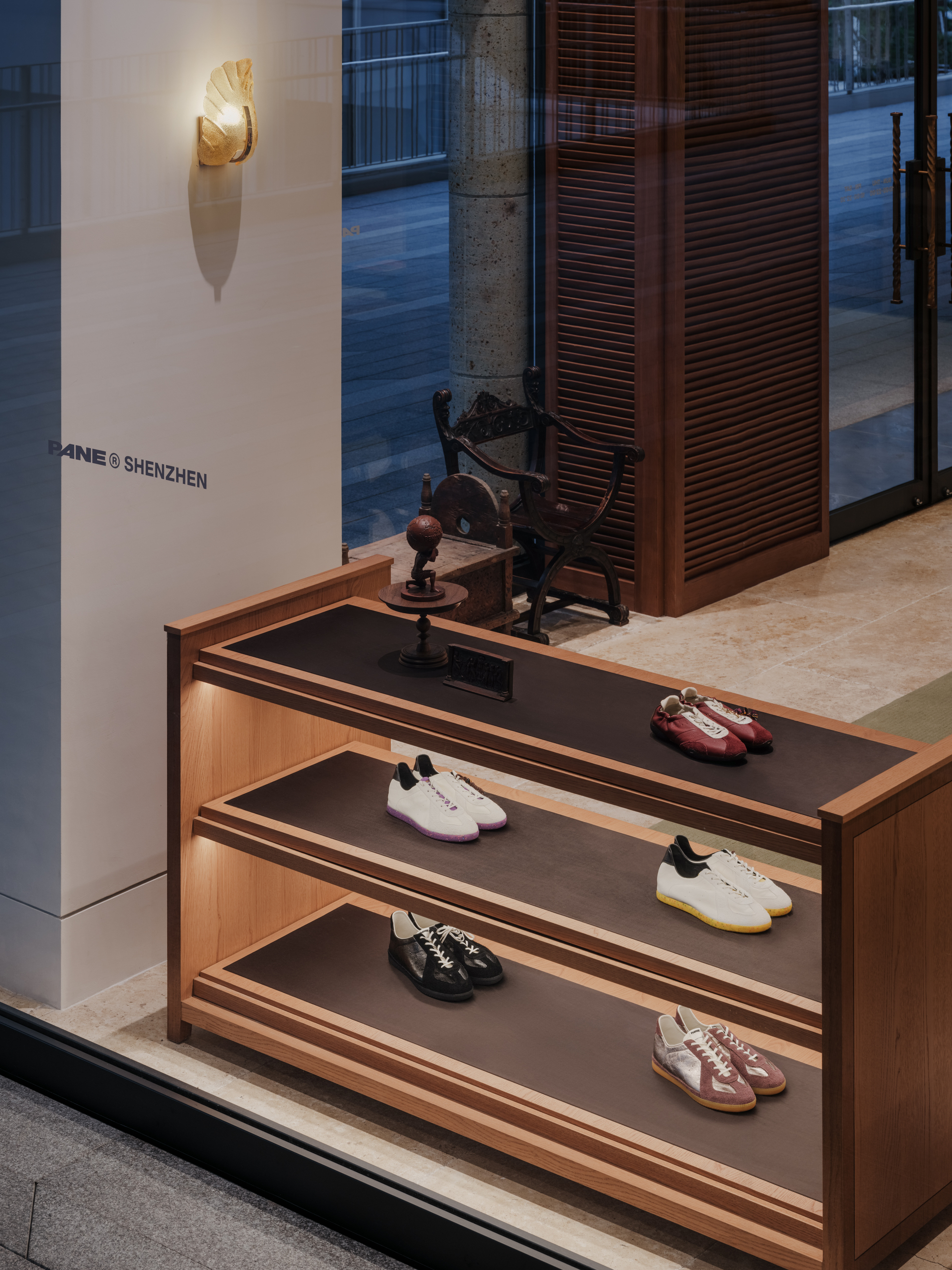 Three-tier timber display shelf of sneakers seen through the shopfront glass at dusk, with a carved chair behind