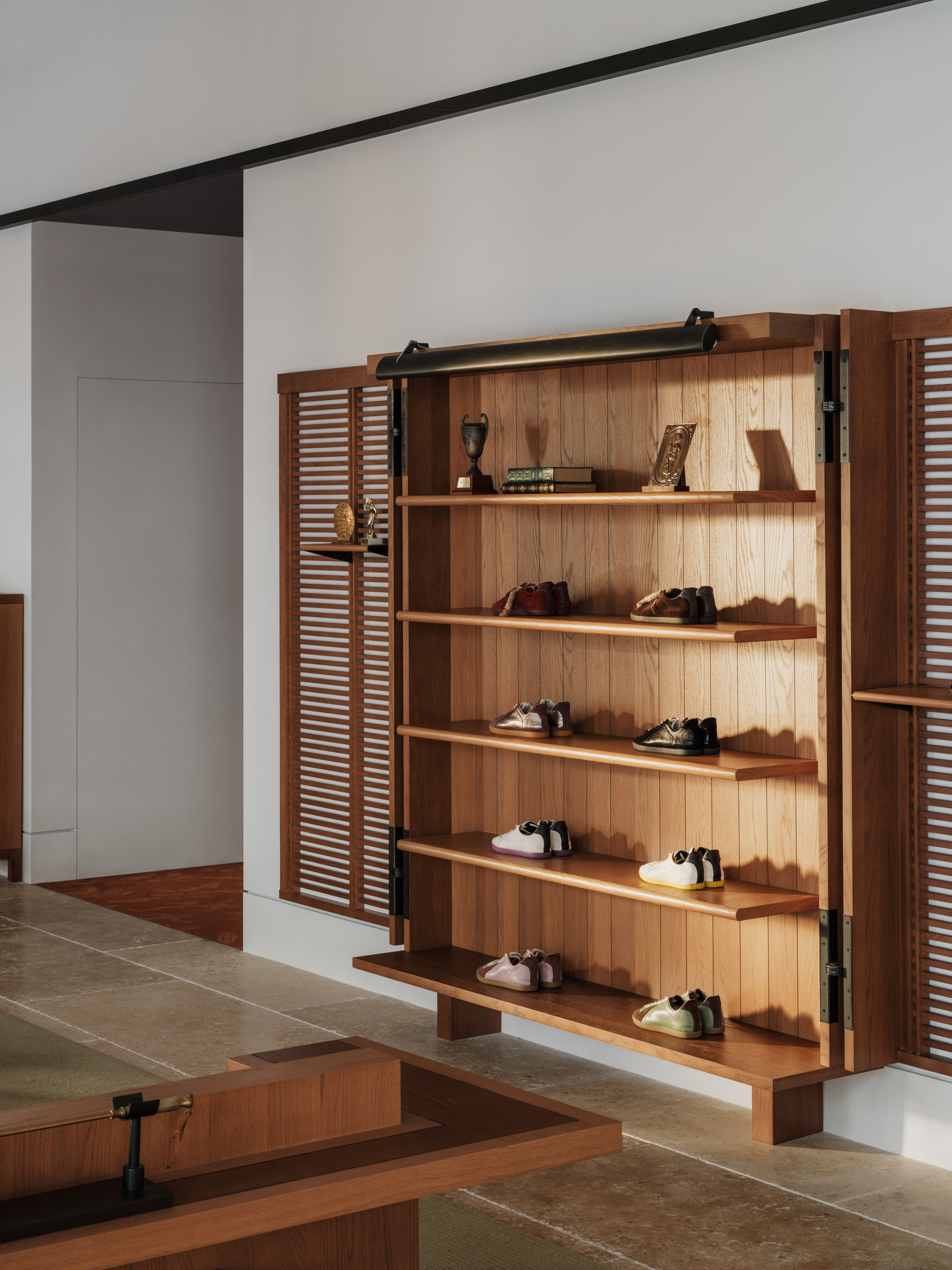 Timber shelving unit with louvred doors and black hinges holding shoes and trophies, lit by warm low sunlight on a stone floor