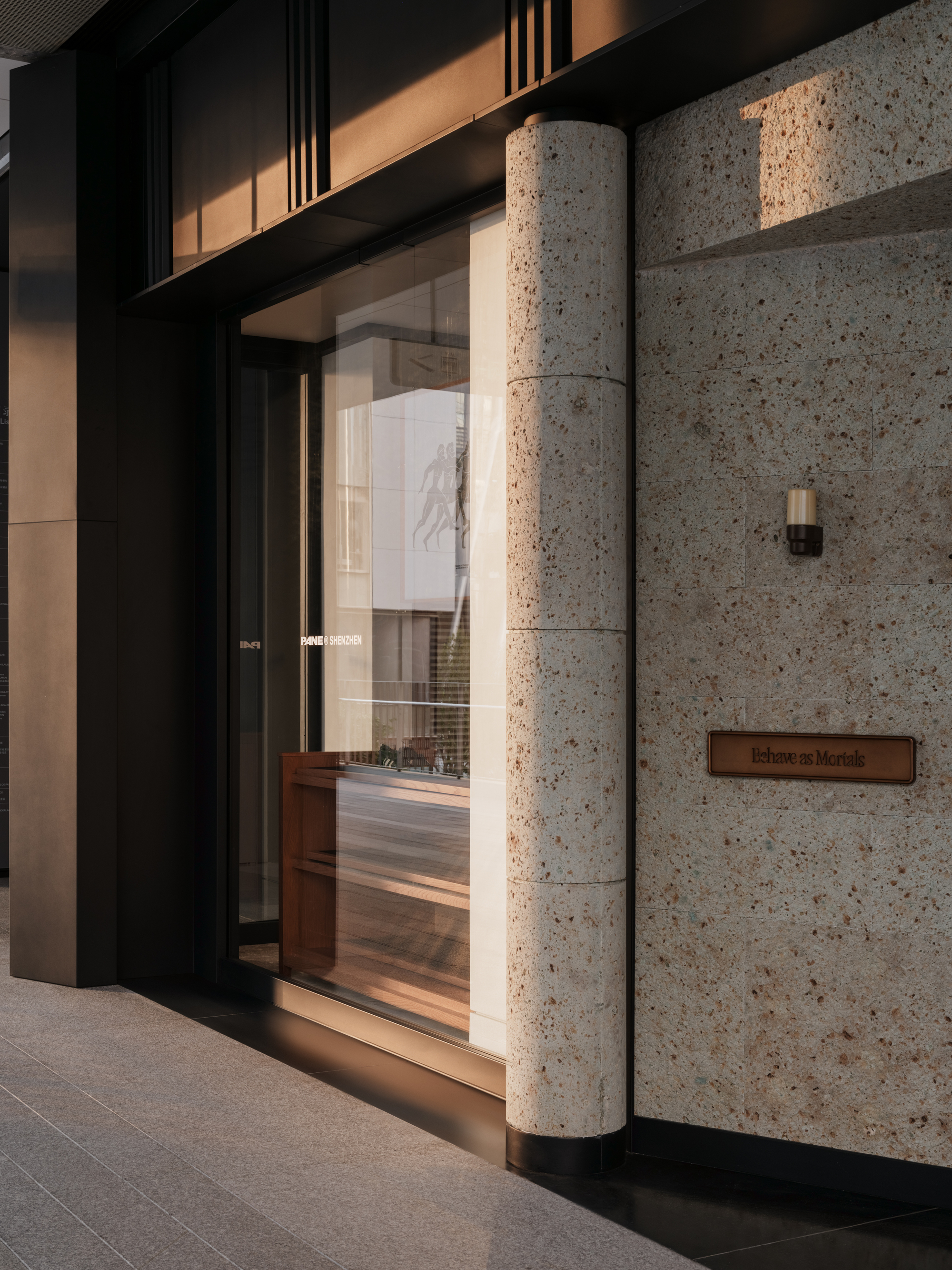 Speckled stone column and wall beside a glazed shopfront with bronze-toned panels, lit by low warm sunlight
