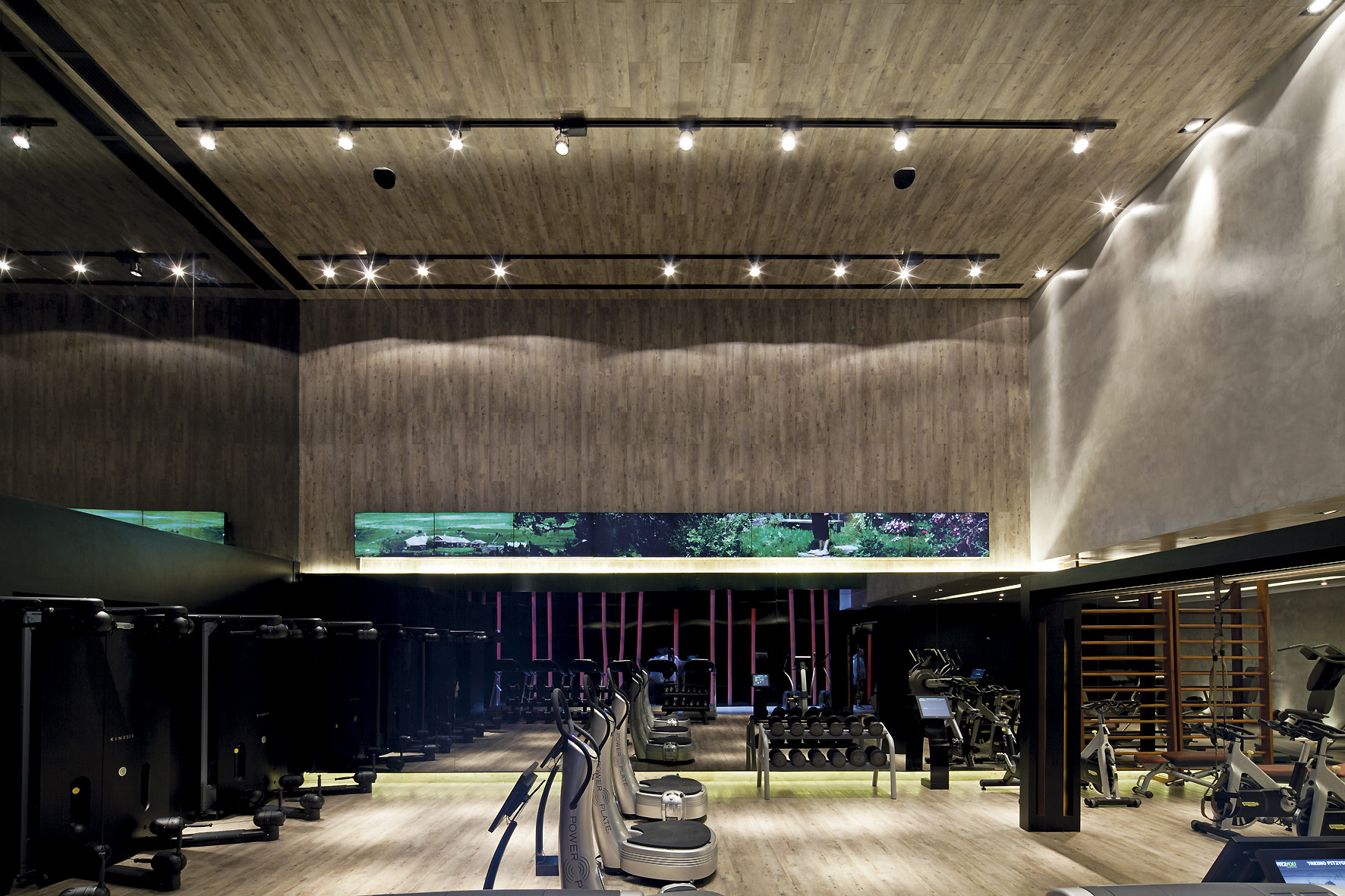 Double-height gym hall with timber-clad walls and ceiling, track lights and a long green video band above rows of black machines