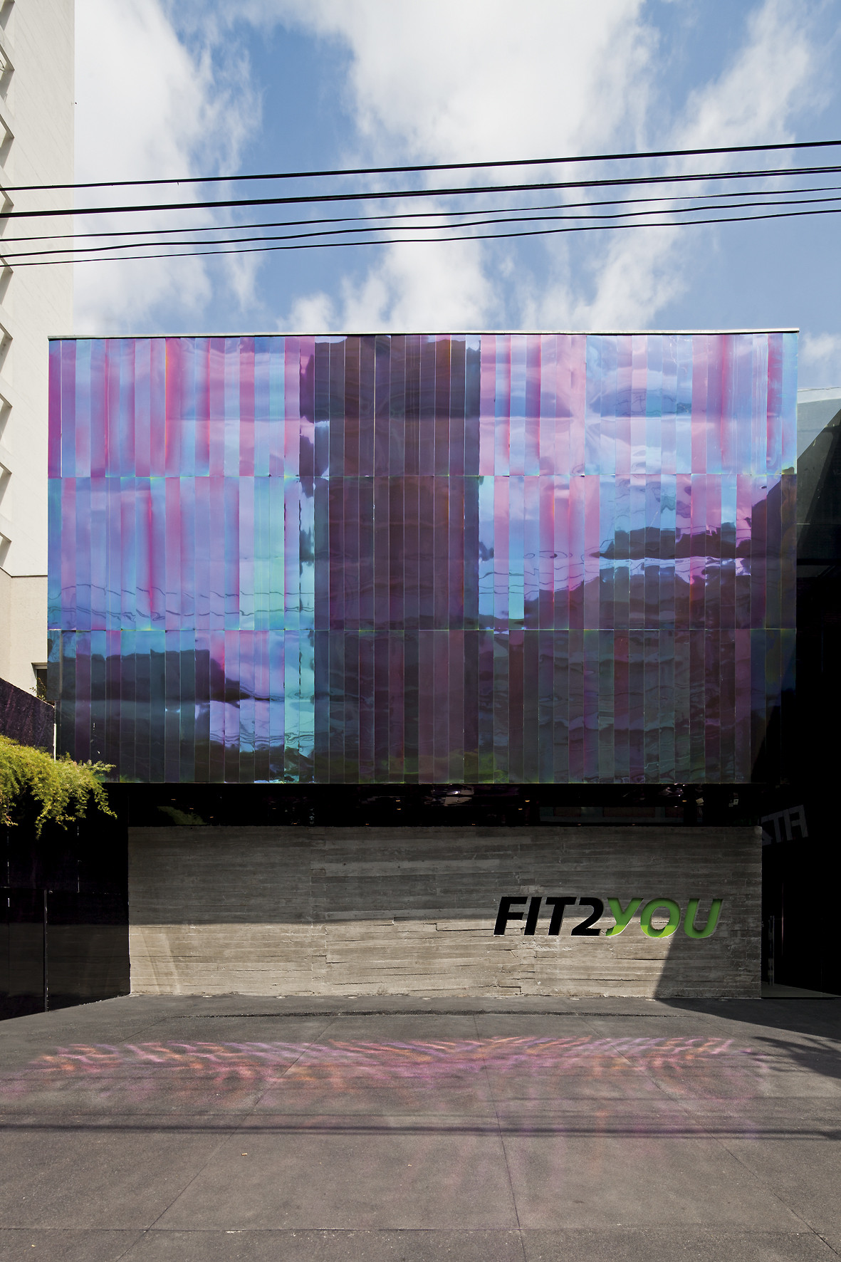 Street facade with an iridescent pink and teal glass screen above a board-formed concrete base under a cloudy blue sky