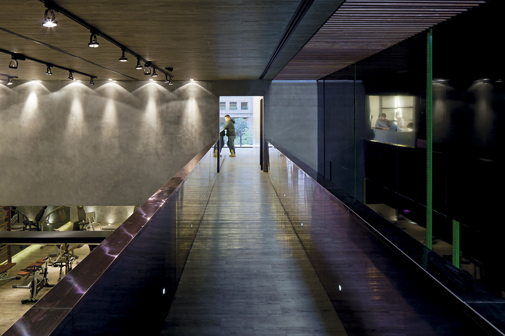 Glass-balustraded bridge with timber flooring crossing above the gym floor, a person walking toward daylight at the far end
