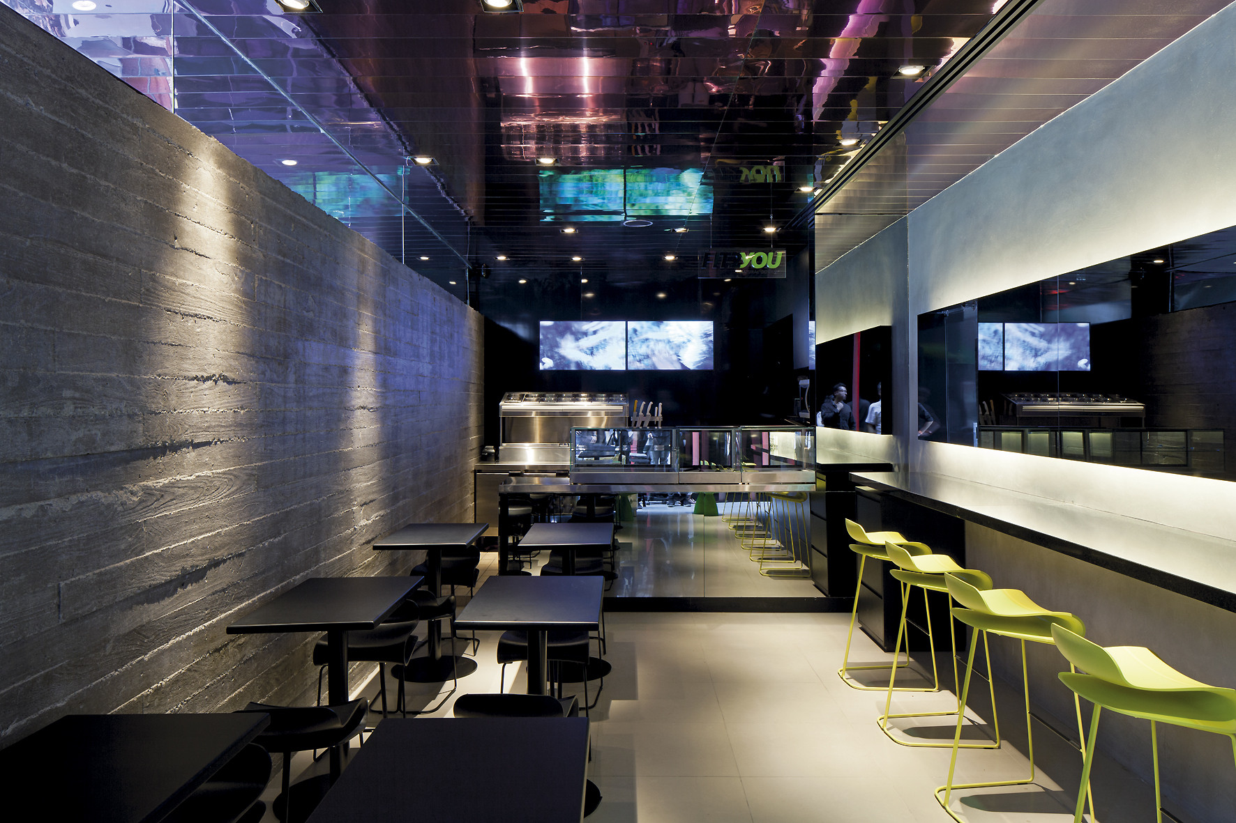 Cafe counter with lime green stools beside a board-formed concrete wall, mirrored ceiling and screens glowing in purple light
