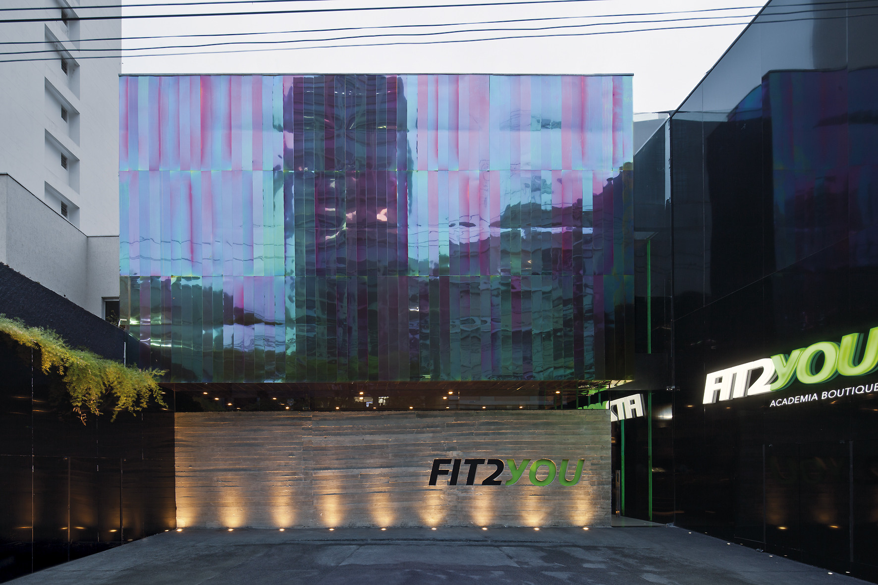 Dusk view of the iridescent glass facade over a concrete wall washed by ground lights, with illuminated signage on the right