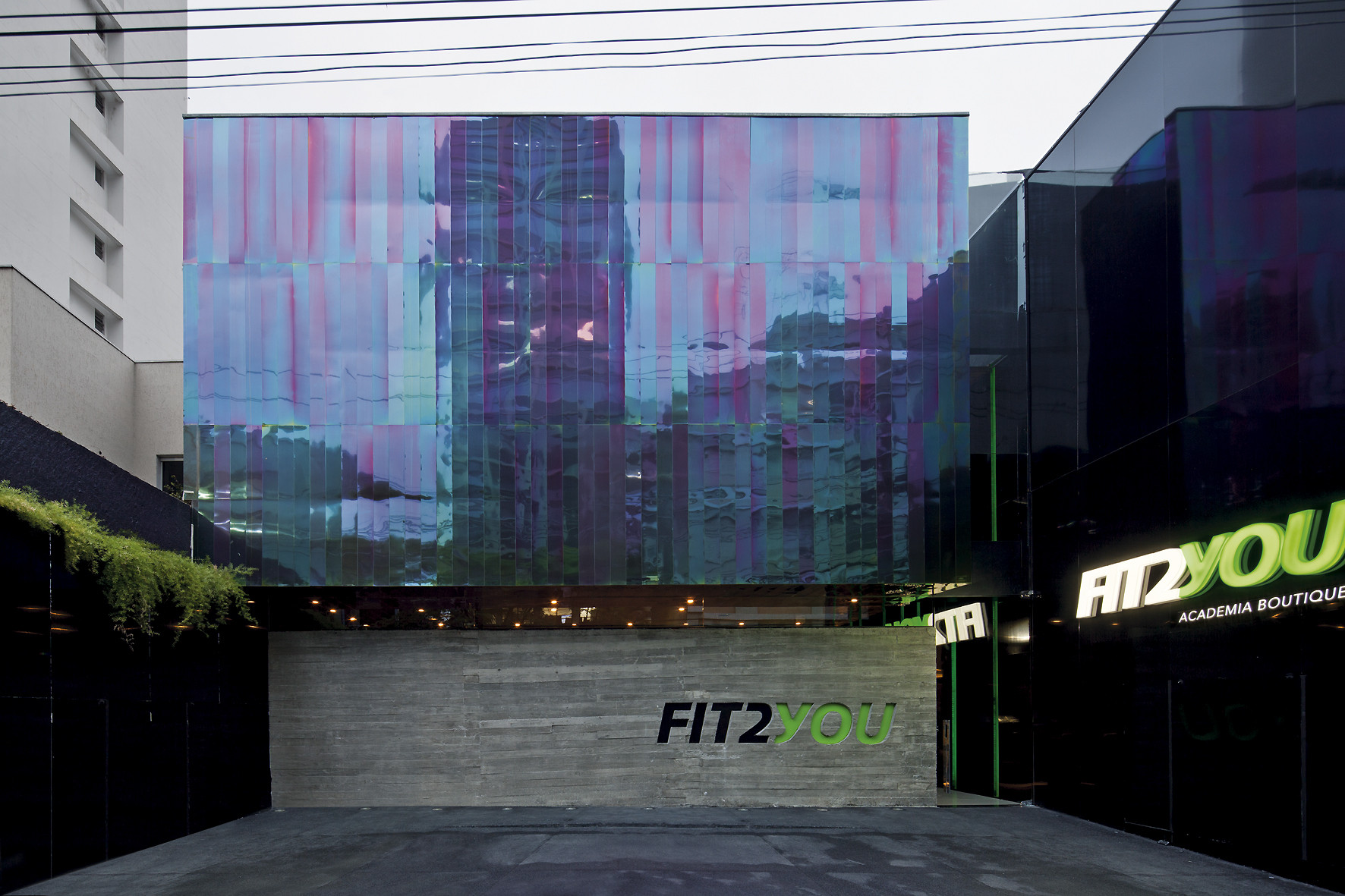 Iridescent pink and blue glass facade above a board-marked concrete wall with green lettering, seen under an overcast sky