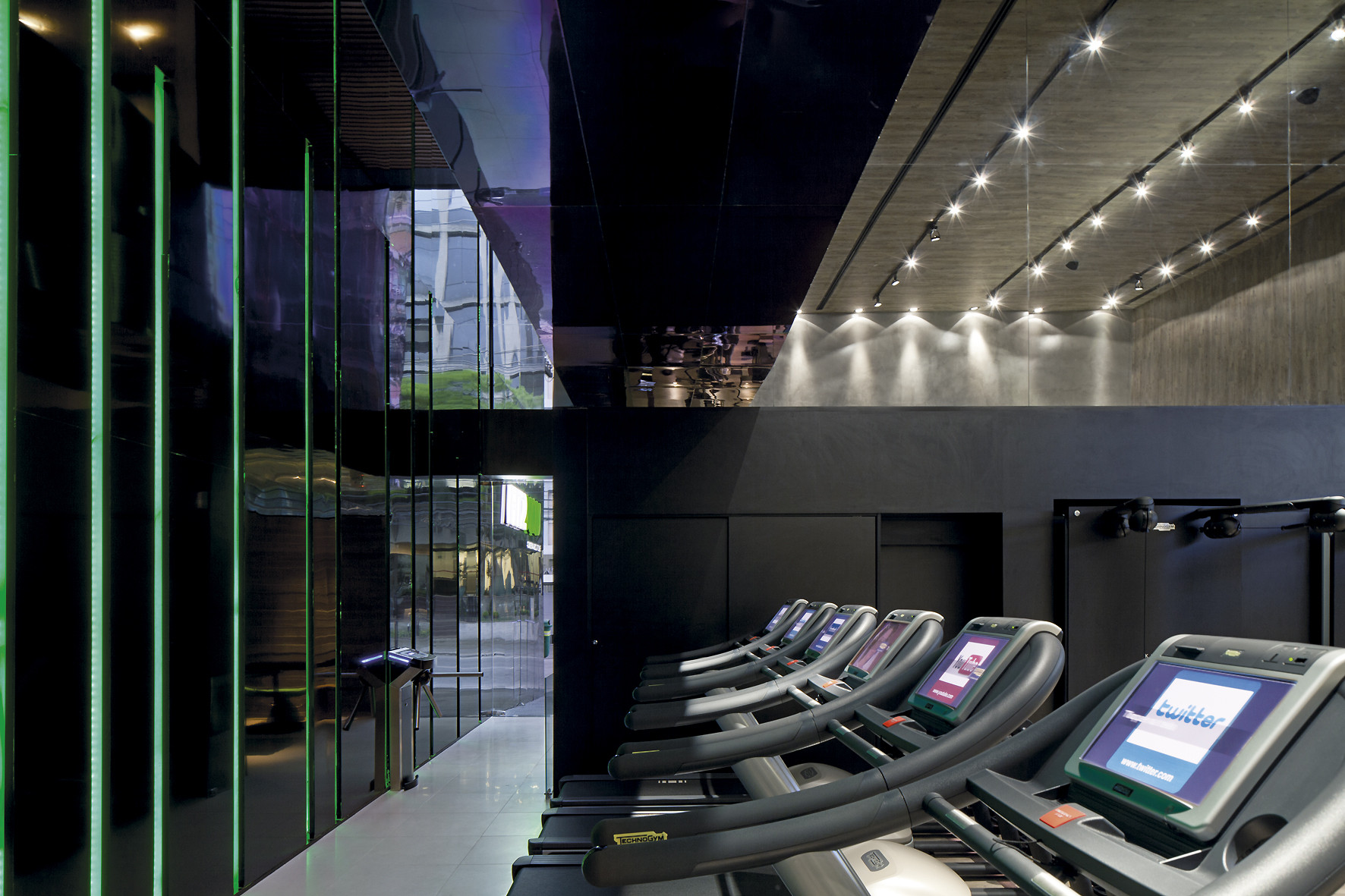 Row of treadmills facing a black mirrored wall edged with green vertical lines, with spotlights washing the concrete ceiling