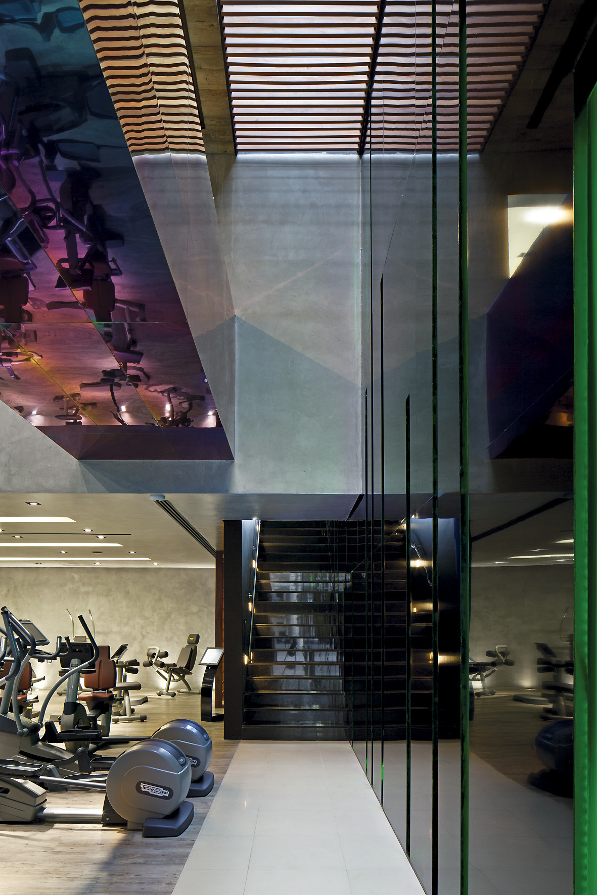 Vertical view of a dark staircase and cardio machines beneath a mirrored ceiling recess and slatted timber skylight