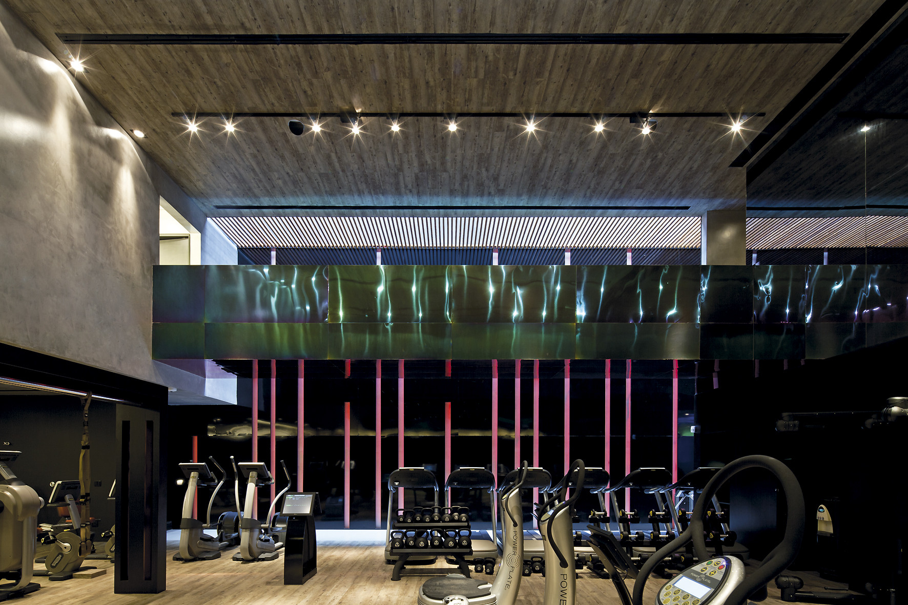 Training floor with cycles and weight racks below a green iridescent metal band and pink vertical rods at dusk